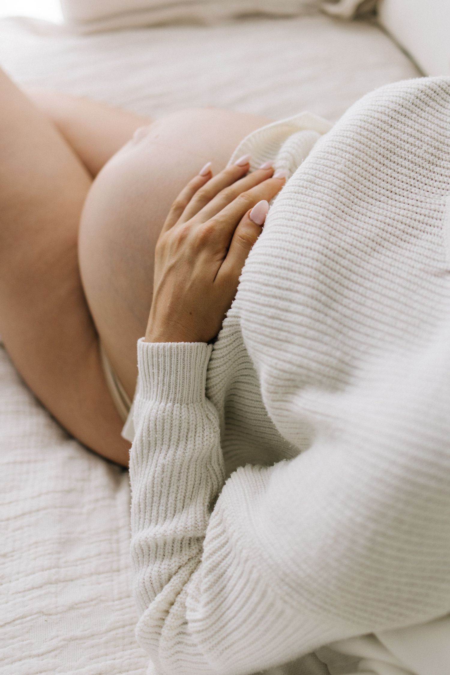 Pregnant woman in white knit sweater gently cradling her baby bump while lying on a bed.