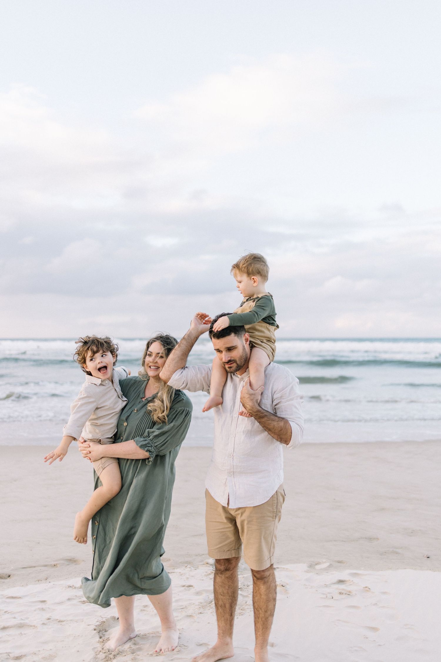 Happy family of four enjoying a beach day, with mom holding a laughing child and dad giving a boy a shoulder ride.