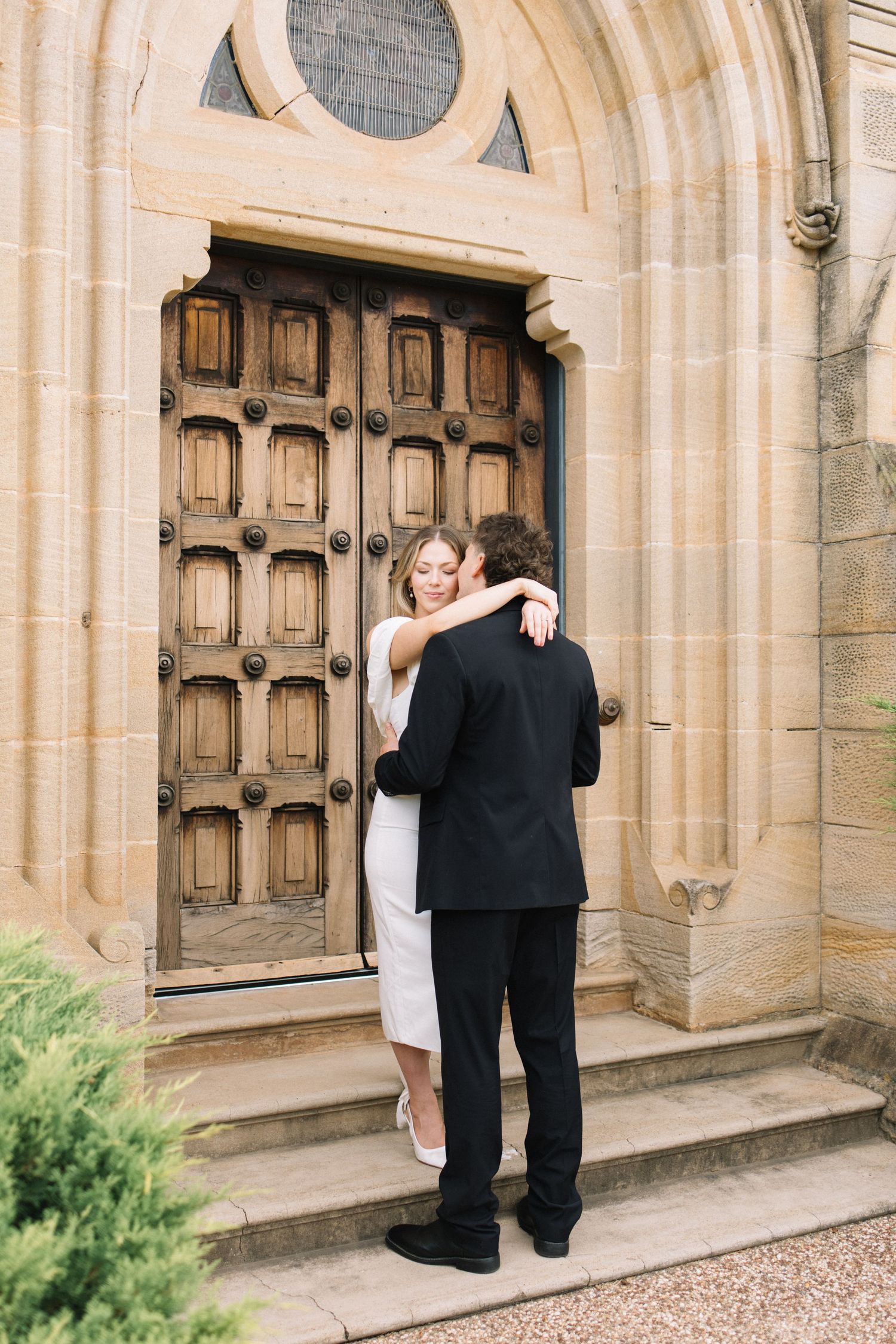 A couple embraces in front of an ornate wooden church door, dressed in wedding attire on stone steps.