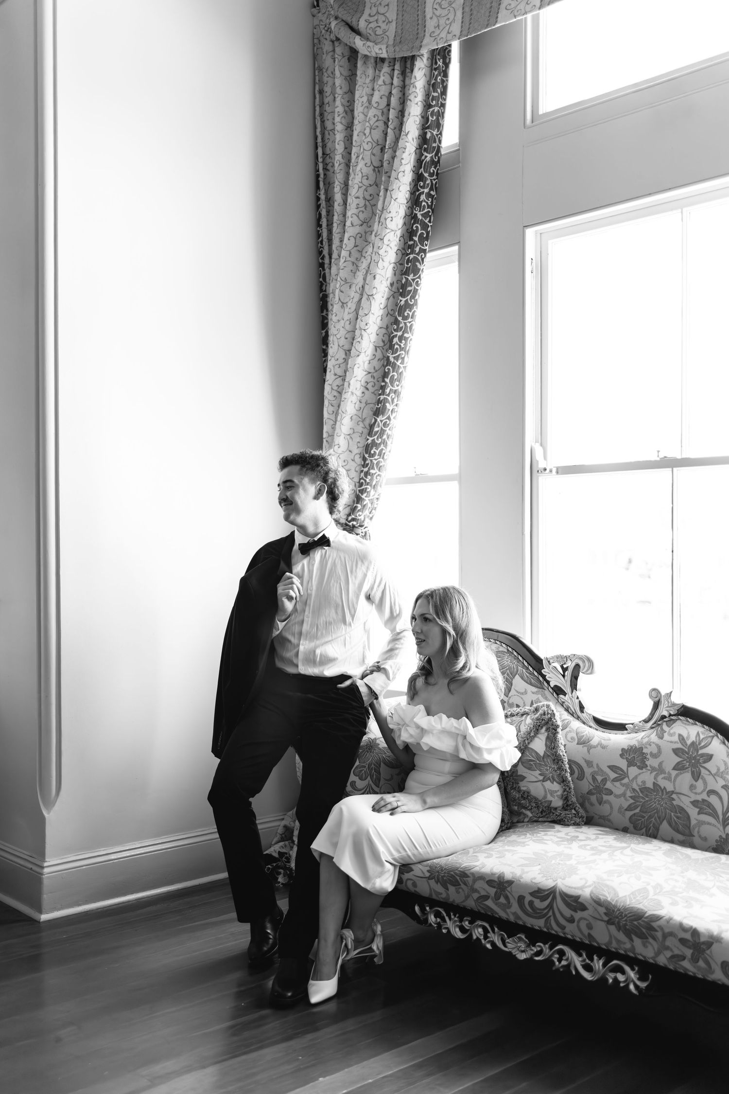 Black and white photo of elegant couple, groom in tuxedo standing by window, bride in white dress seated on ornate sofa.
