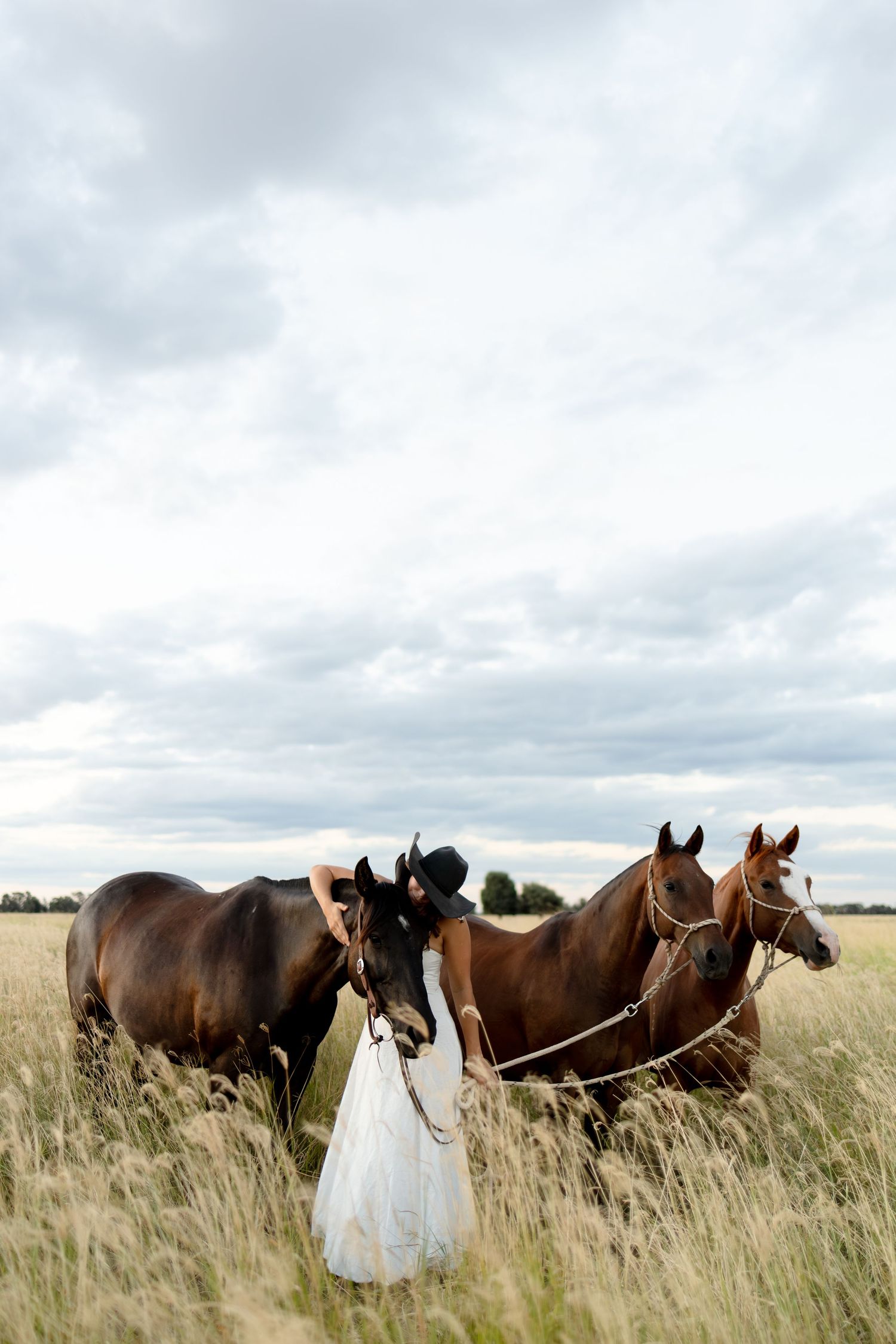 Woman in white dress and black hat standing with three brown horses in a vast open field.