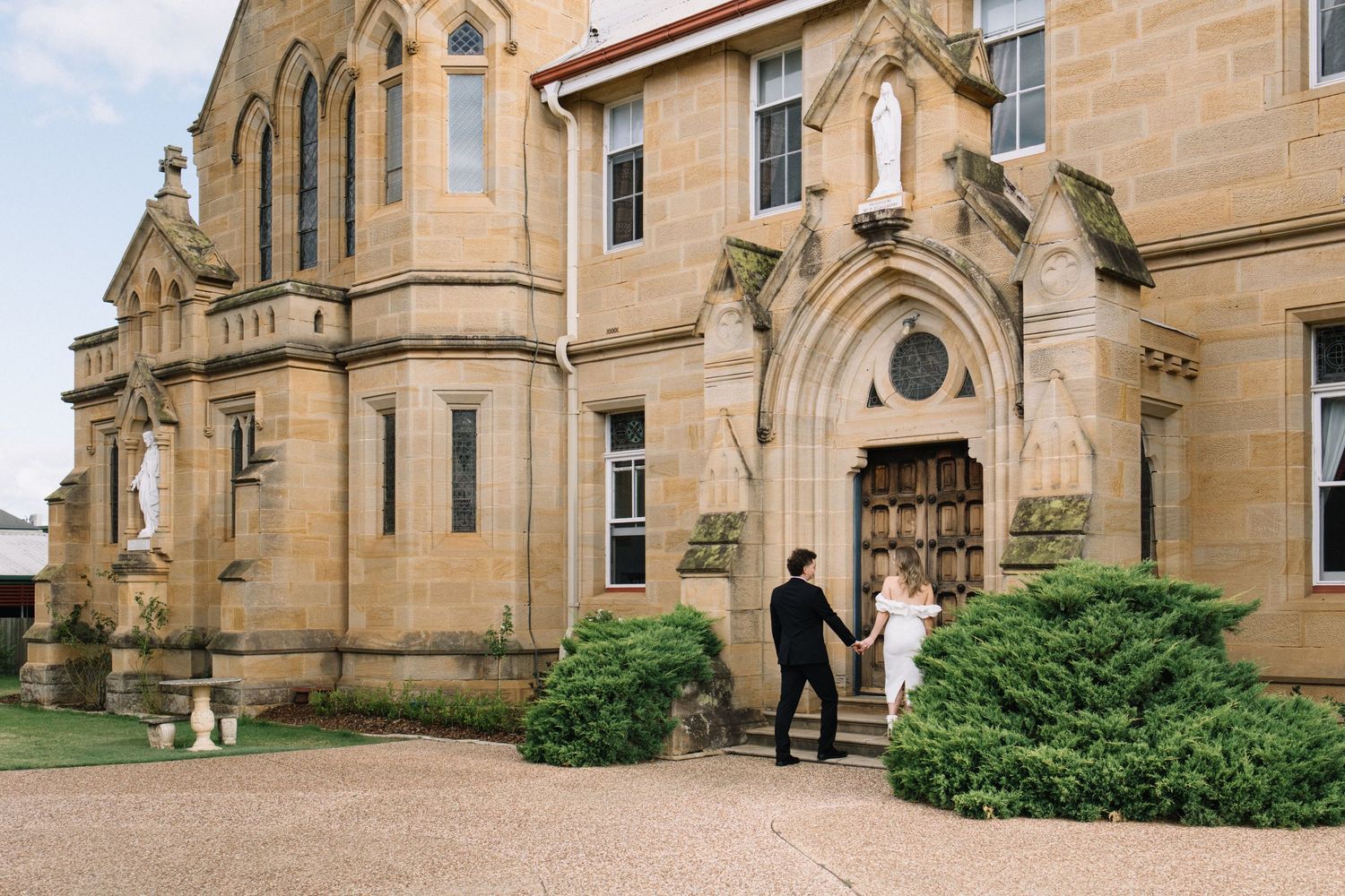 Couple holding hands walking toward ornate sandstone church entrance on their wedding day.
