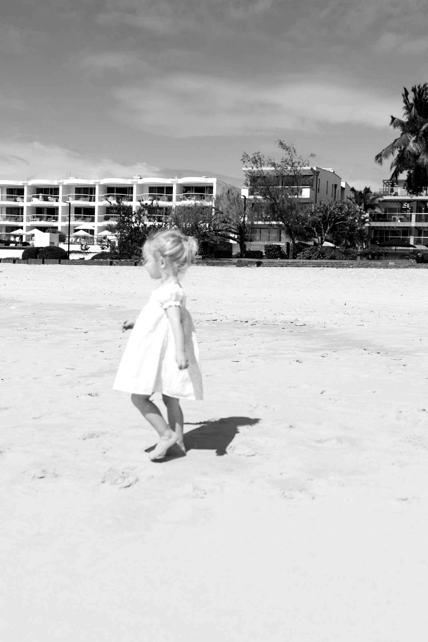 Black and white photo of a young blonde girl in a white dress walking barefoot on a sandy beach near a resort.