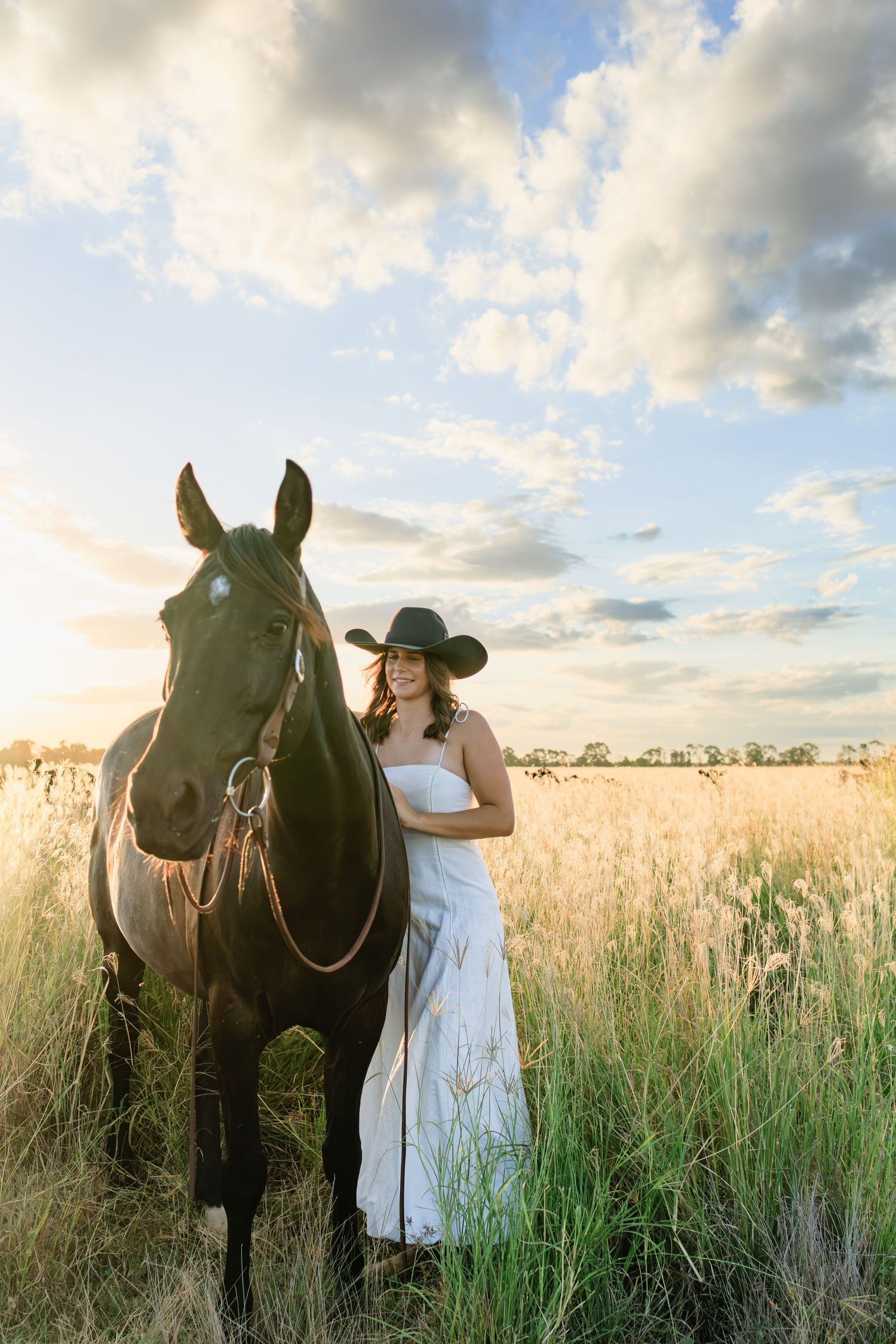 Smiling woman in white dress and black cowboy hat standing beside a dark horse in golden field.