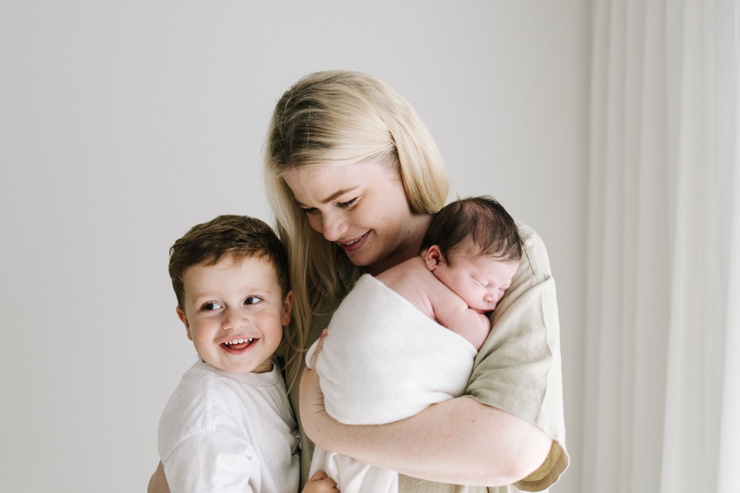 A smiling blonde mother holds her sleeping newborn while her young son smiles beside her.