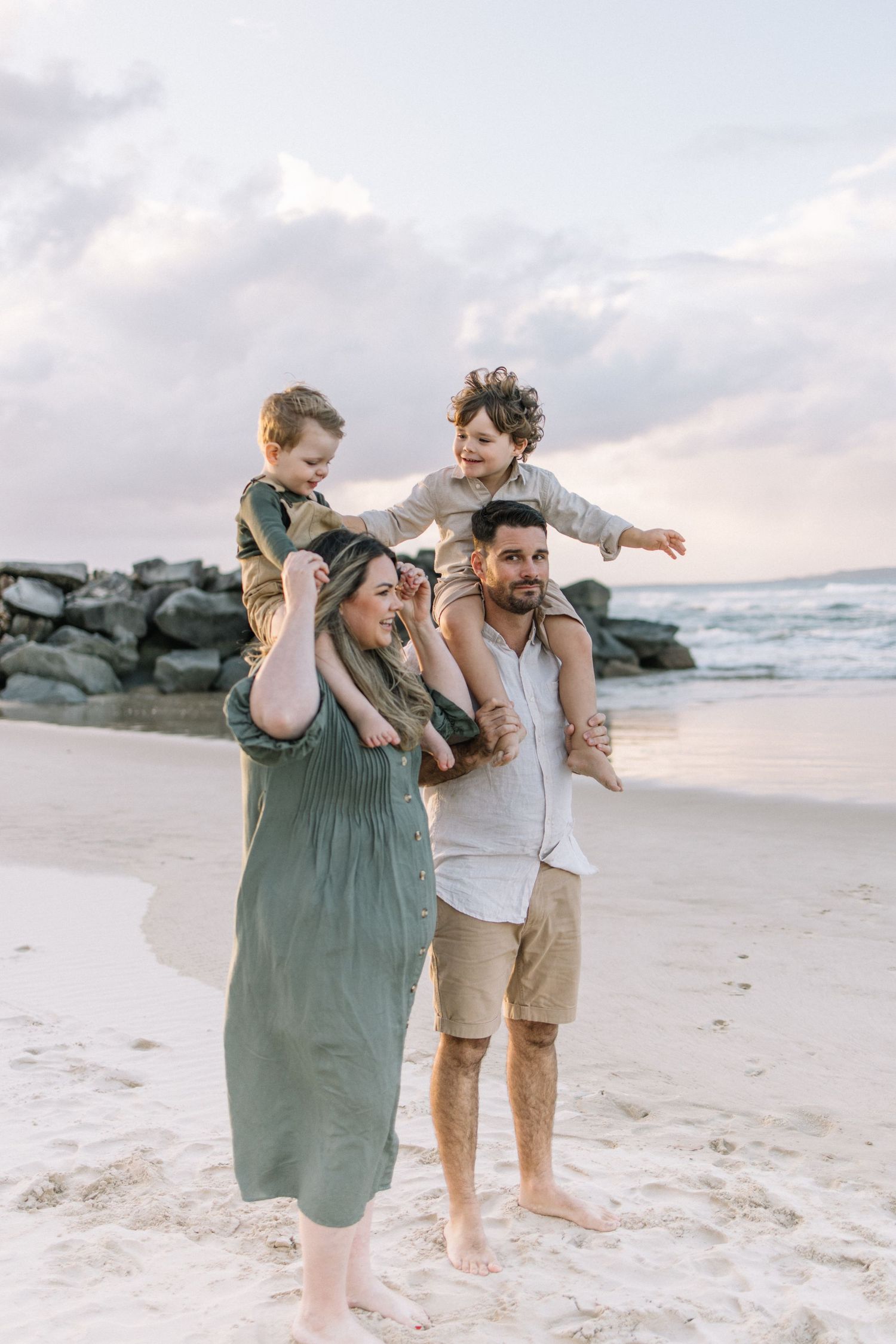 Joyful family of four at the beach, two young boys sitting on parents shoulders at dusk.