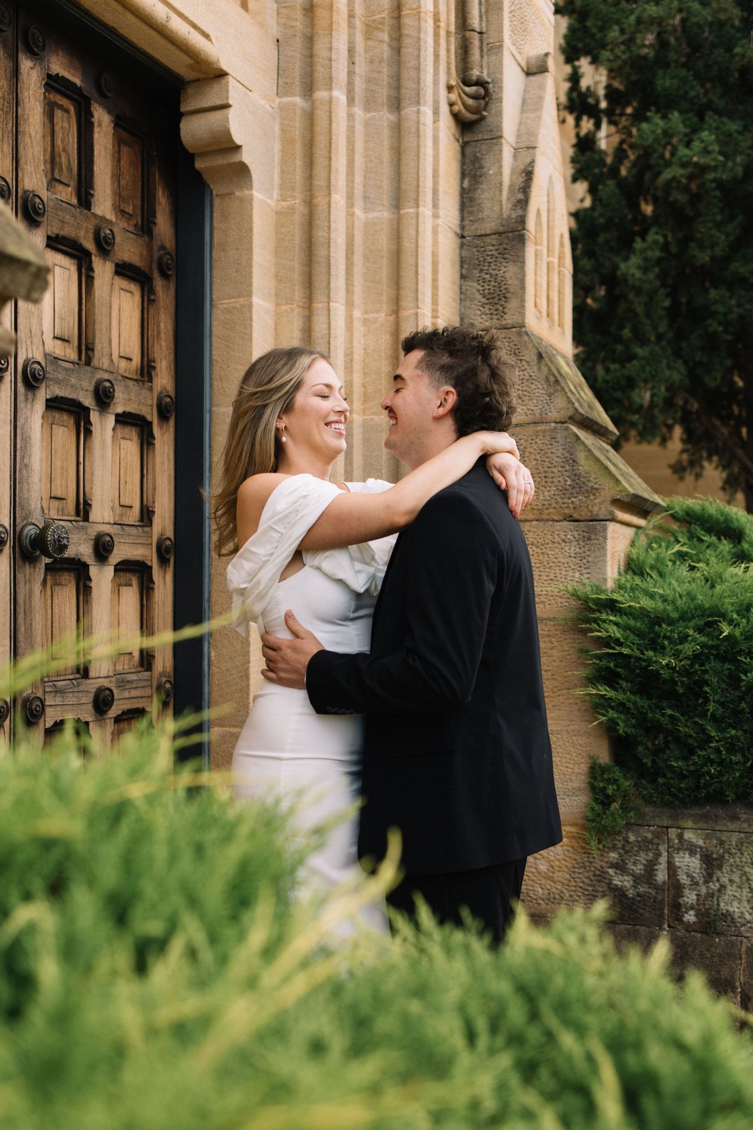 A joyful couple embraces and smiles outside a stone church with wooden doors and green hedges.