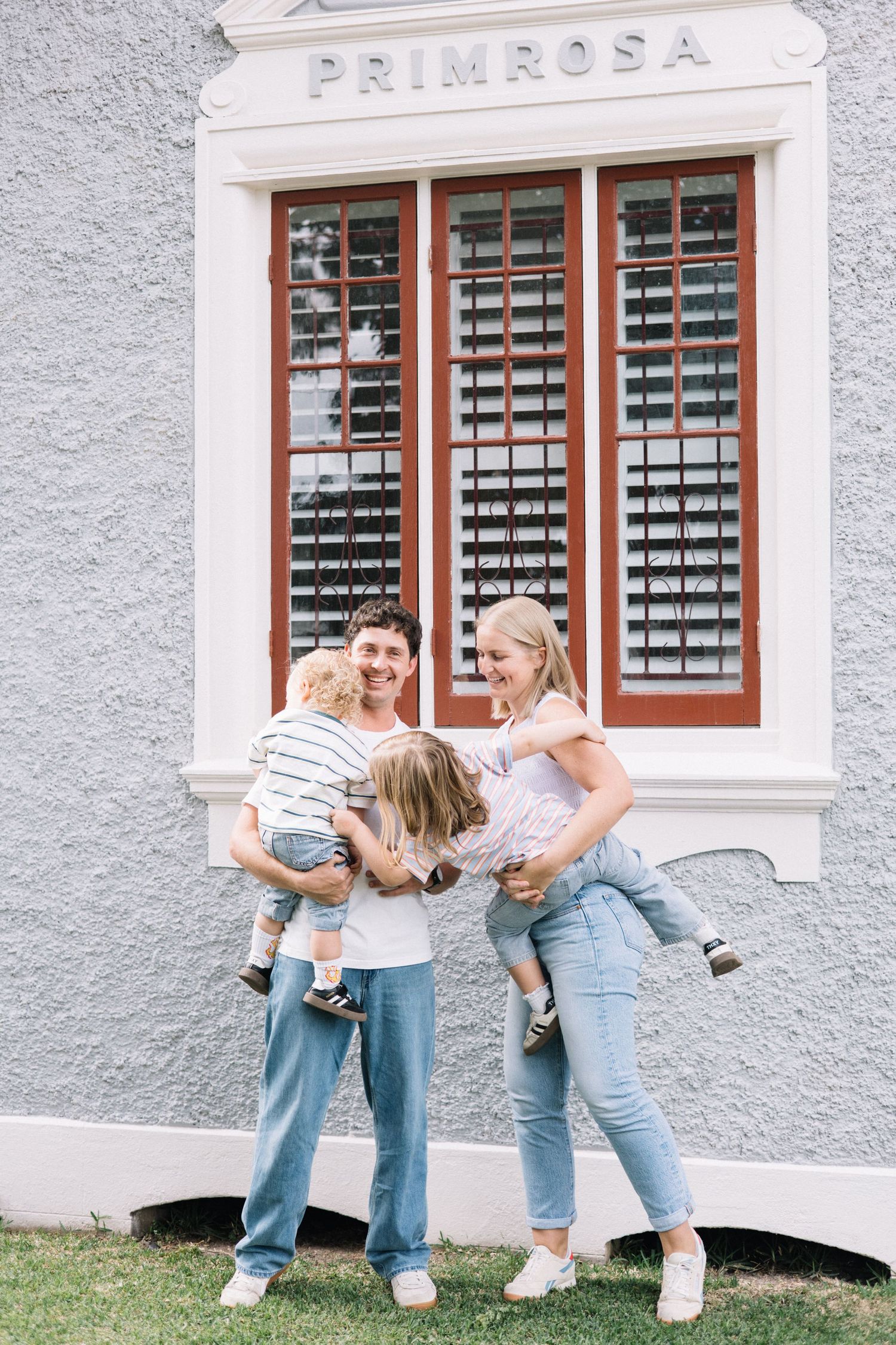 Happy family of four posing outside a building named Primrosa, parents holding two young children, wearing casual denim outfits.