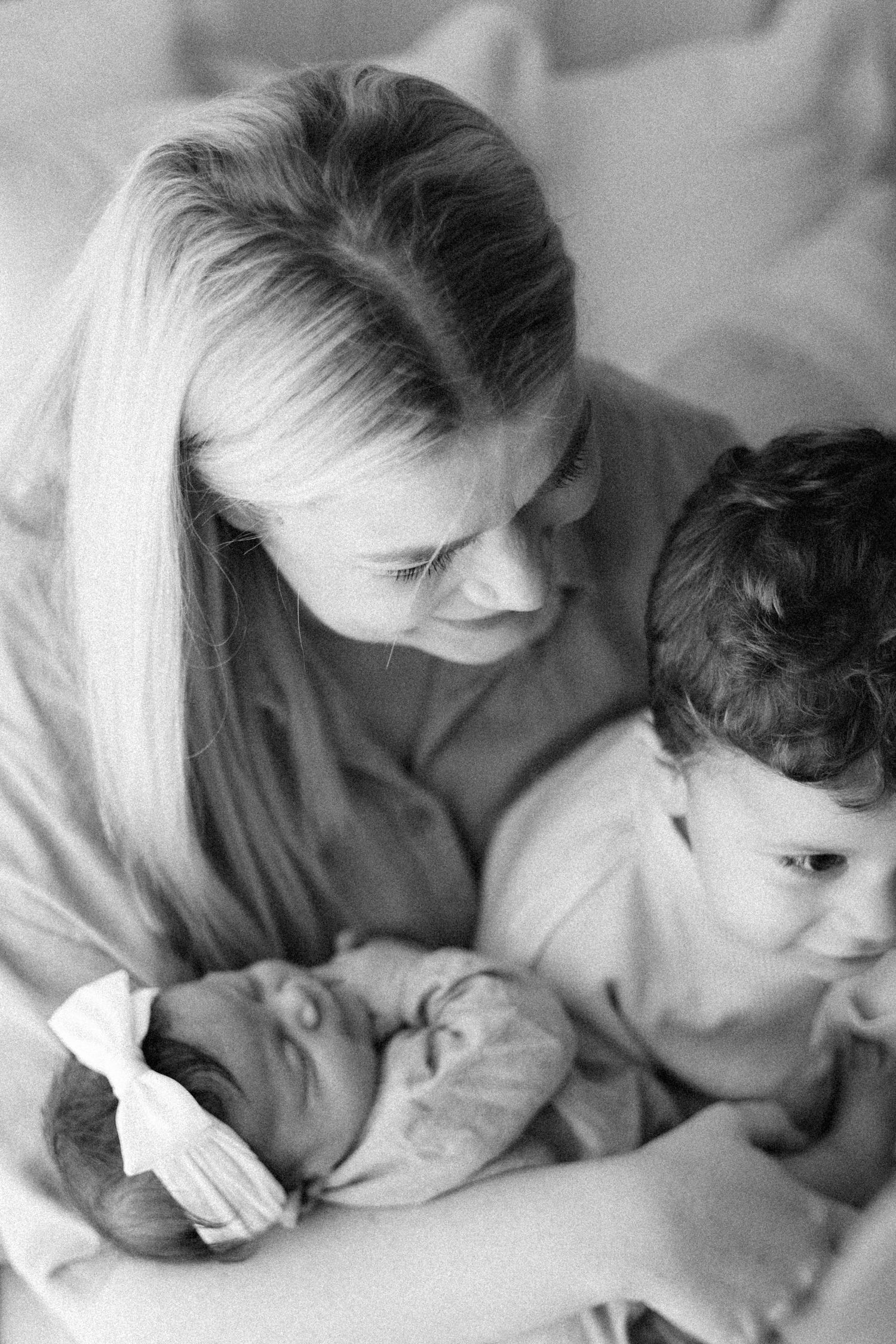 Black and white photo of a blonde mother tenderly holding a newborn baby and young toddler child.