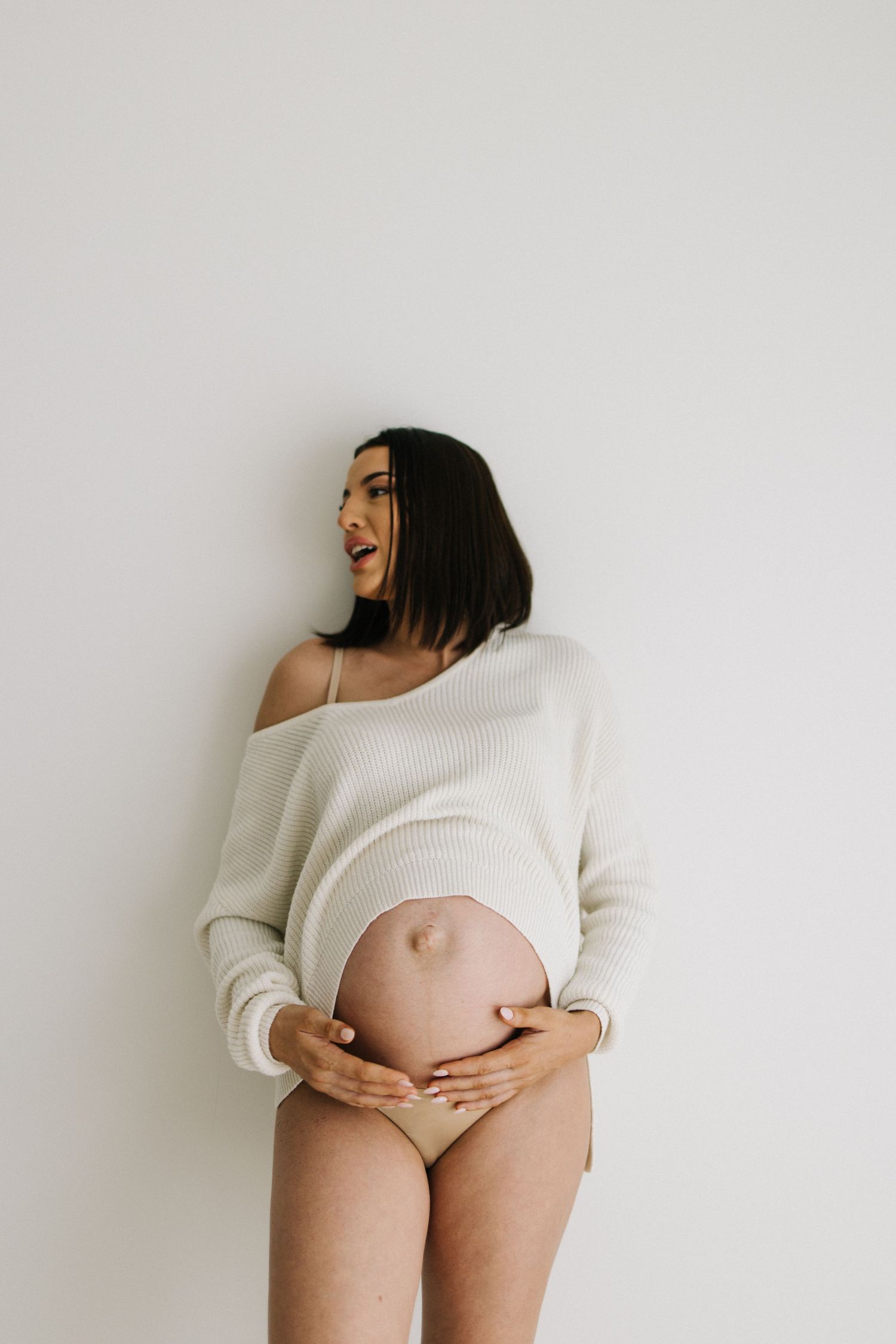 Pregnant woman in cream knit sweater holding bare baby bump while looking sideways against white wall.
