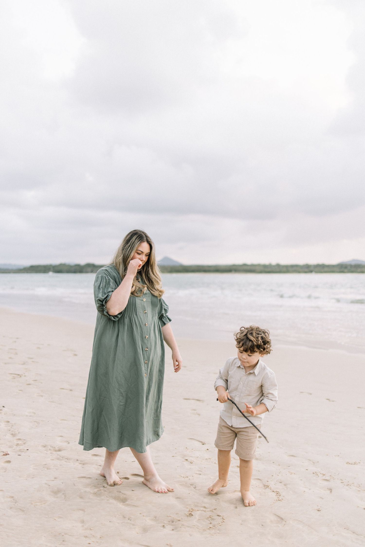 A barefoot mother in a green dress watches her curly-haired son play with a stick on the beach.