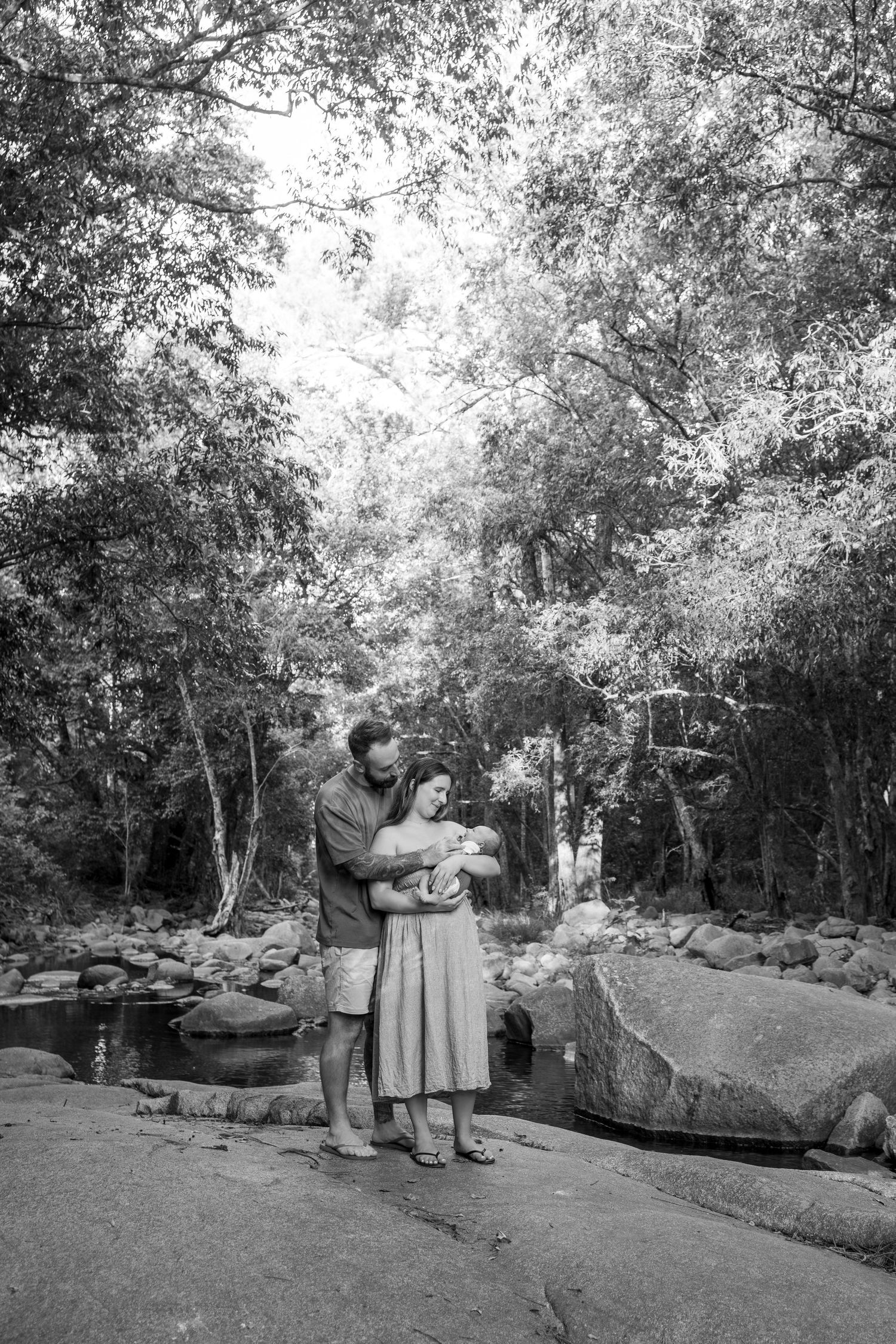 Black and white photo of a couple tenderly holding their newborn baby outdoors near a rocky creek surrounded by lush trees.