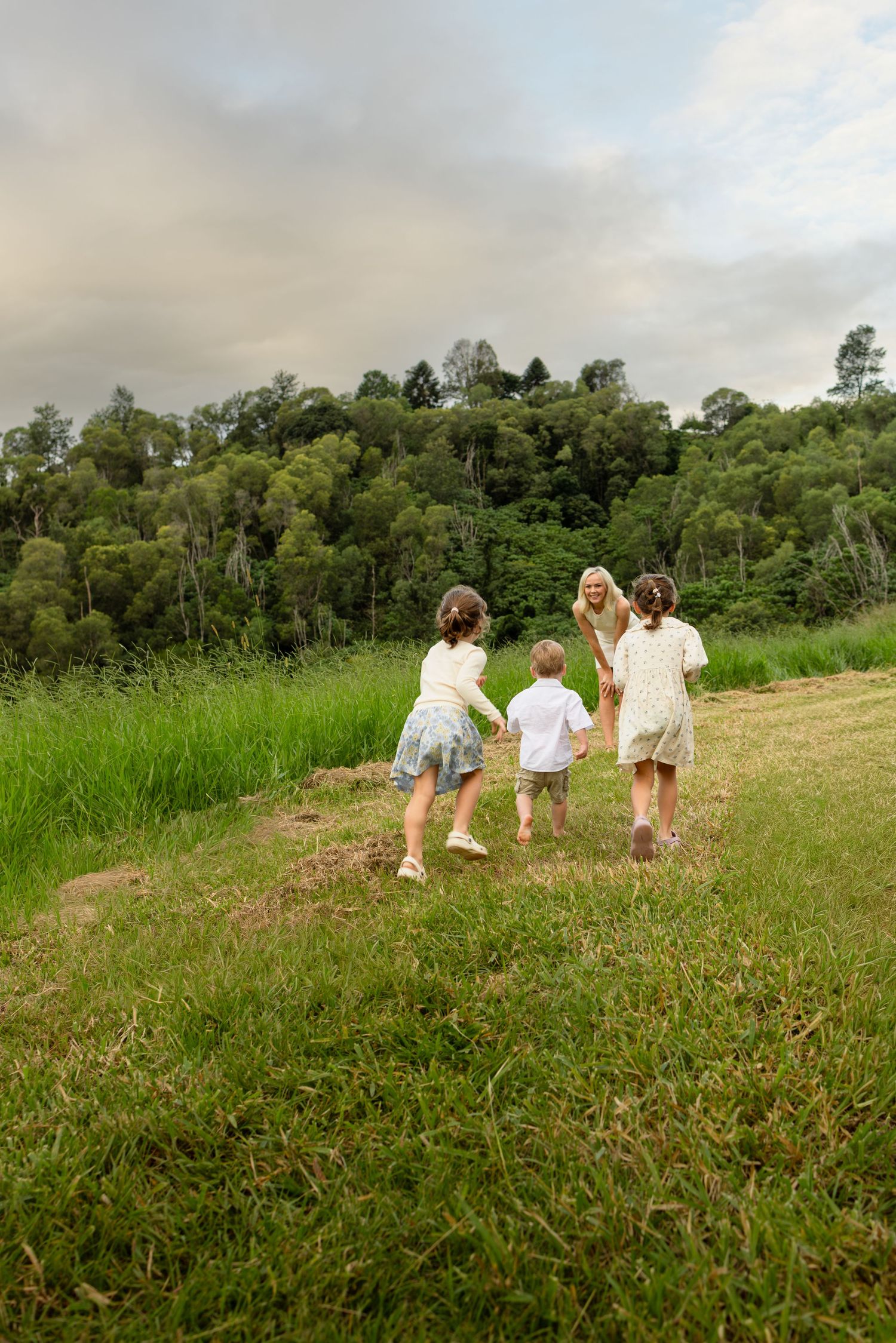 A woman and three children walk hand-in-hand through a lush green meadow toward a dense forest under cloudy skies.