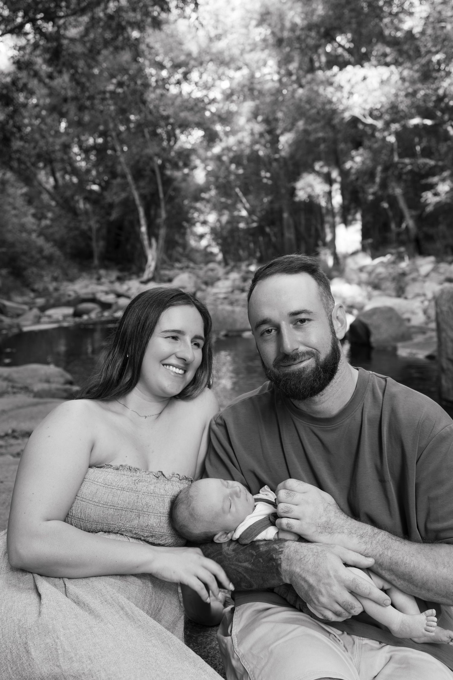 Smiling couple with beards holds newborn baby outdoors near a rocky creek in black and white portrait.