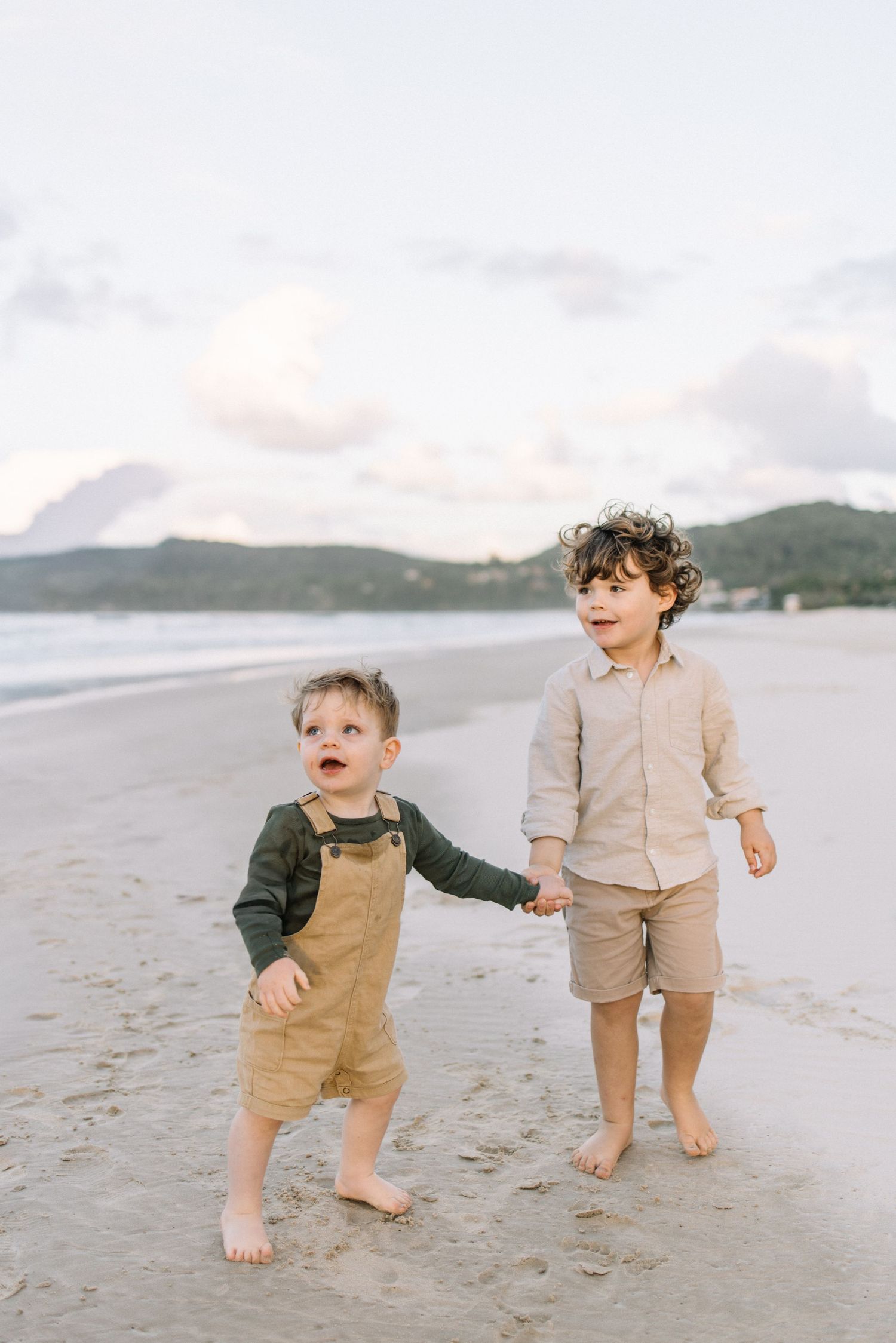 Two young brothers hold hands while walking barefoot on a beach at sunset.