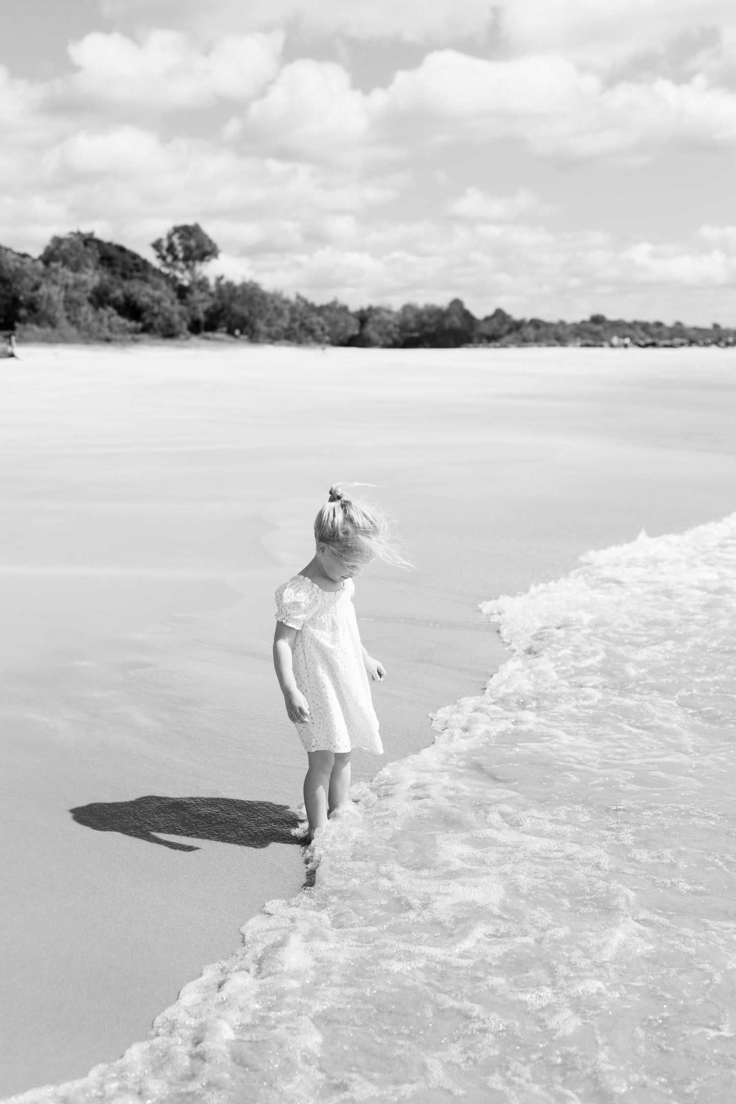 Black and white photo of a young girl in a white dress walking along a sandy beach shoreline with gentle waves.