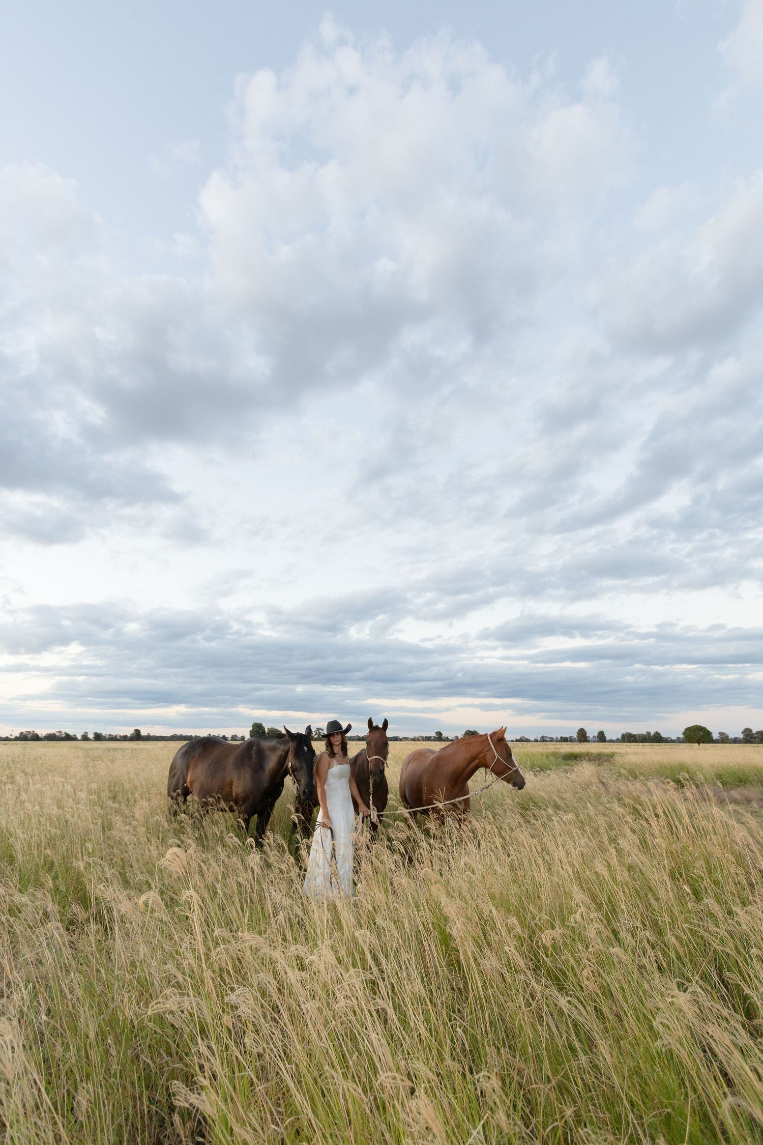 Woman in white dress and cowboy hat leads three horses through tall golden grass under dramatic clouds.