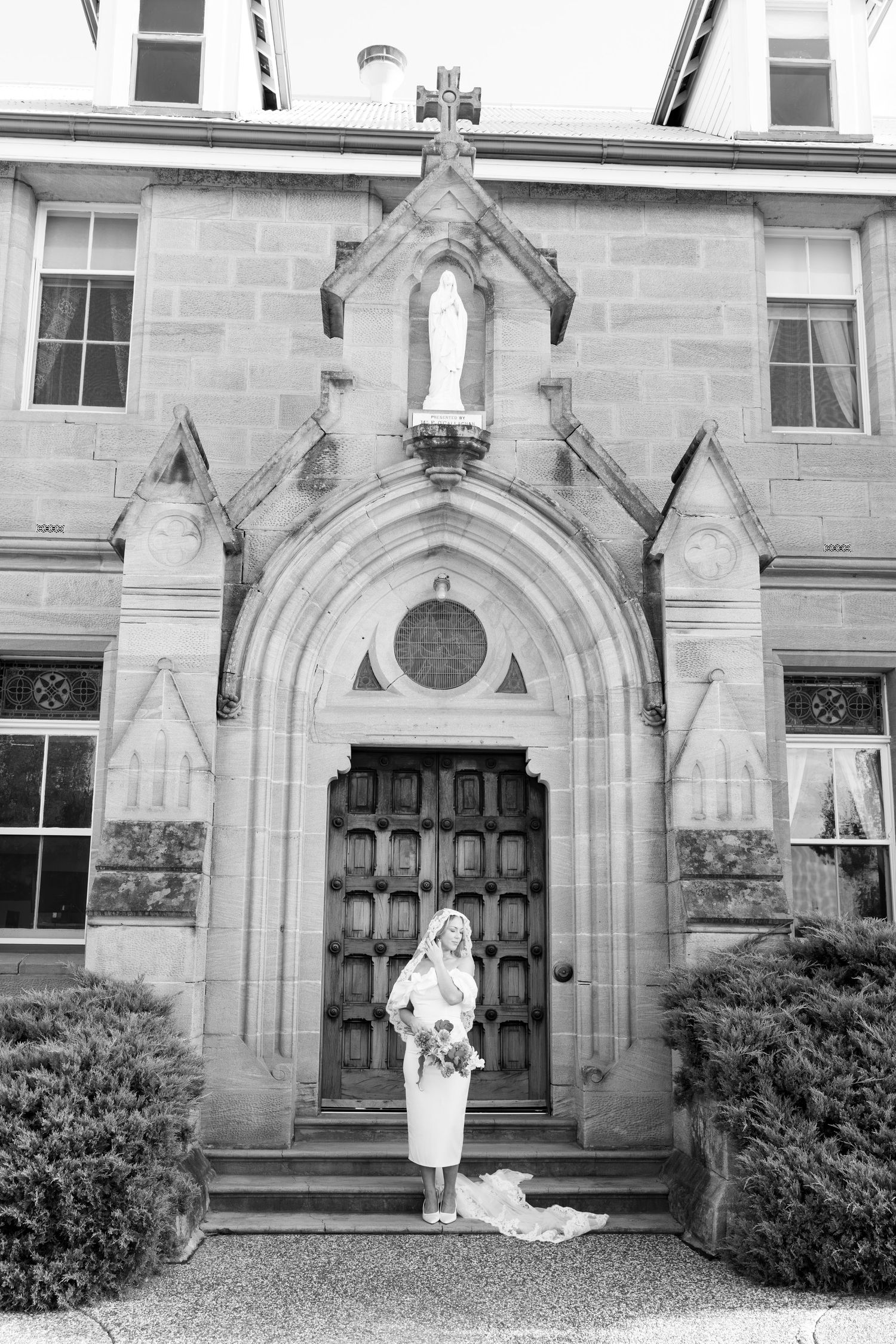 Bride in wedding dress poses before ornate Gothic church entrance with religious statue, black and white photo.