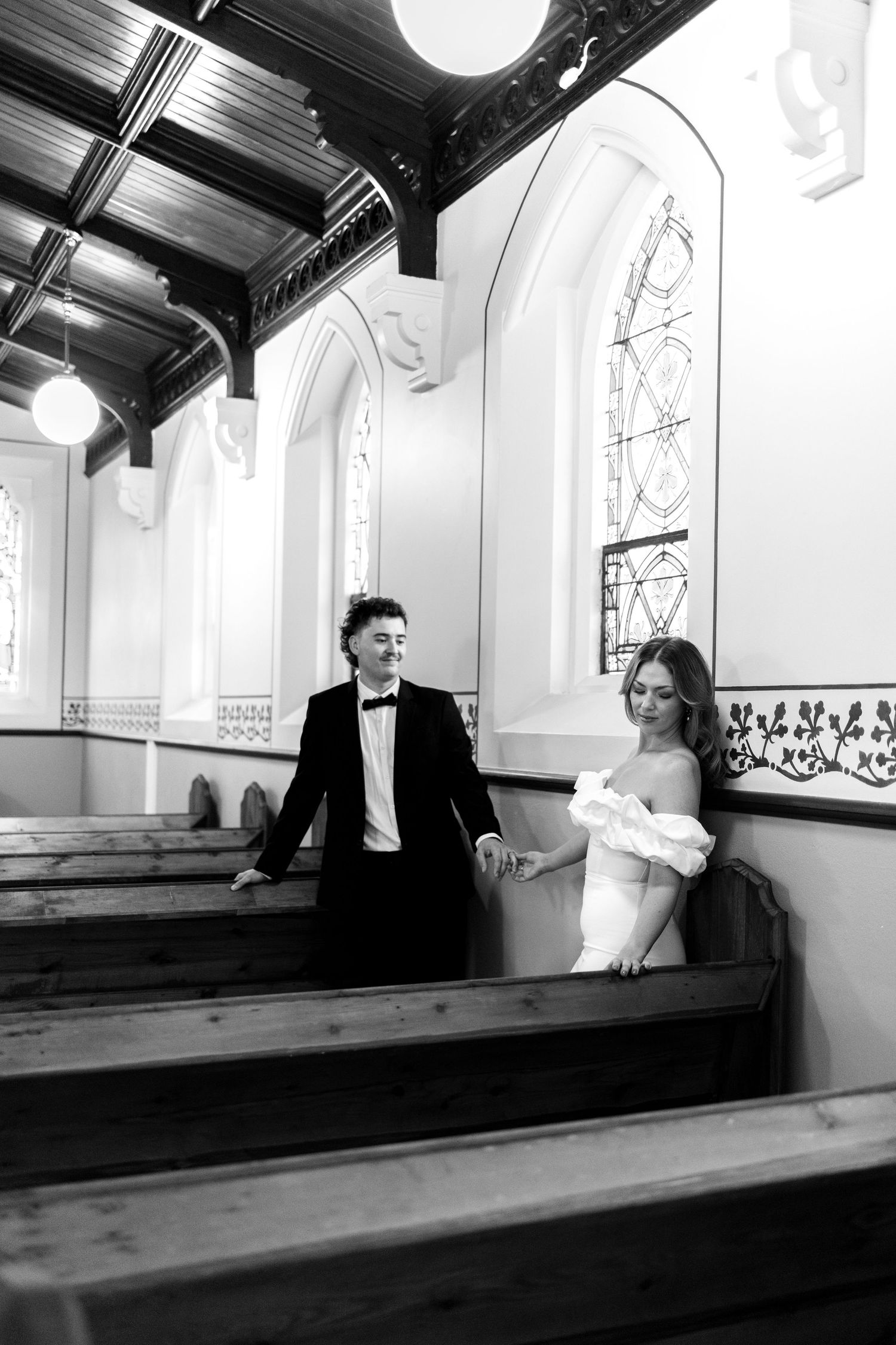 Black and white photo of a bride and groom holding hands in a historic church with wooden pews and stained glass windows.