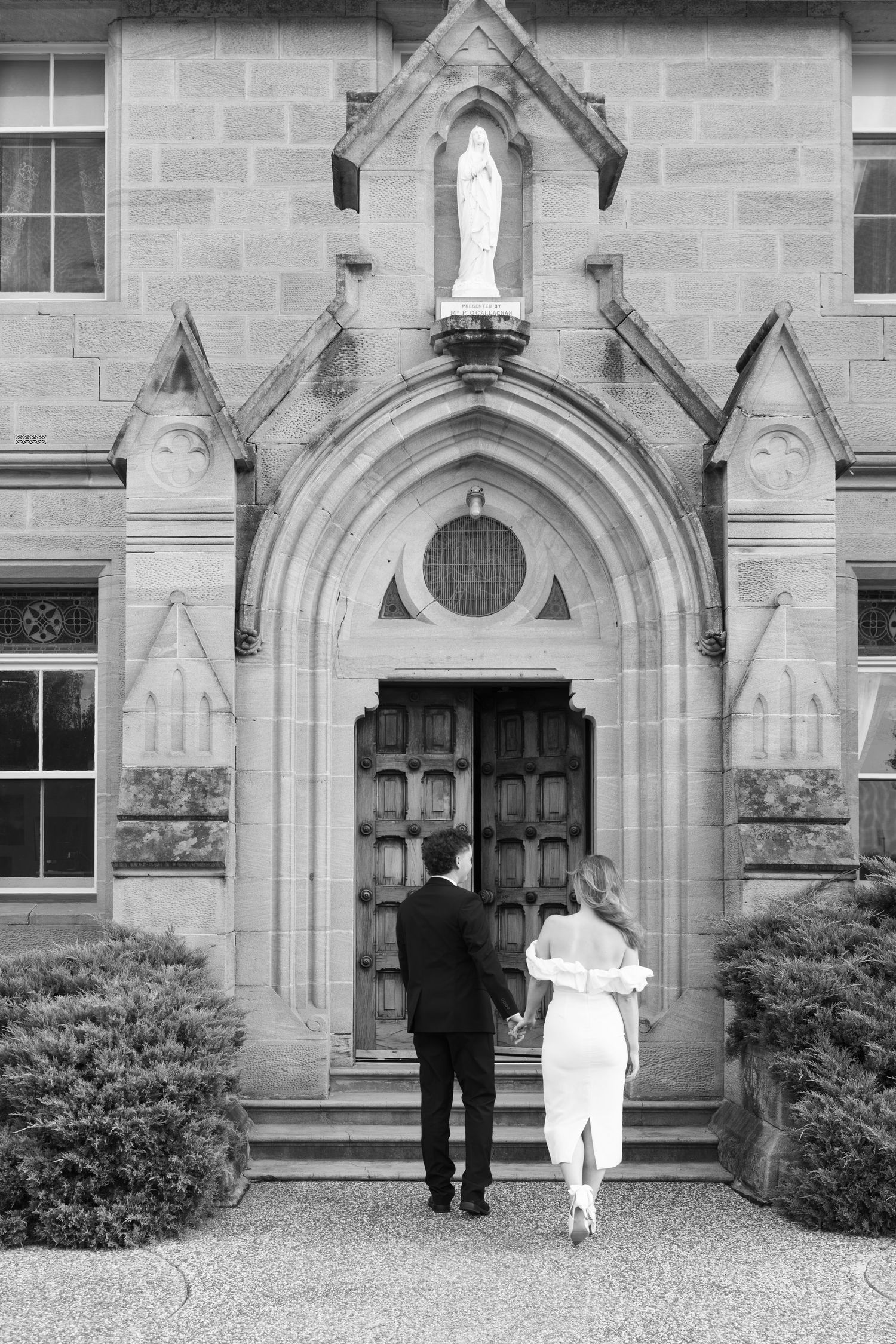 Black and white photo of a bride and groom entering a stone church with Gothic arched doorway and Virgin Mary statue.
