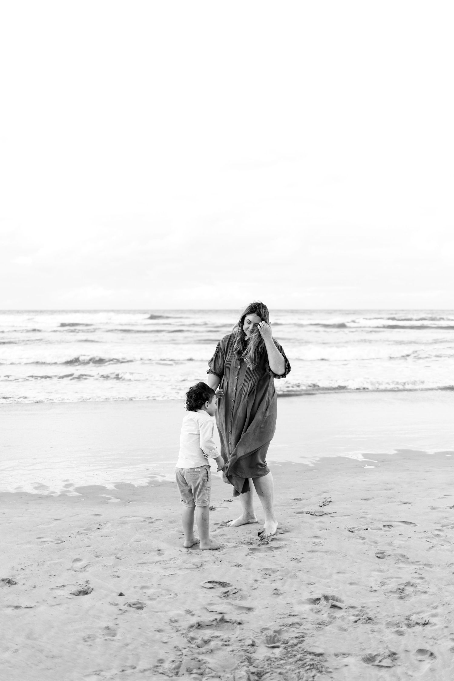 Black and white photo of a mother and young child holding hands on a sandy beach near the ocean.