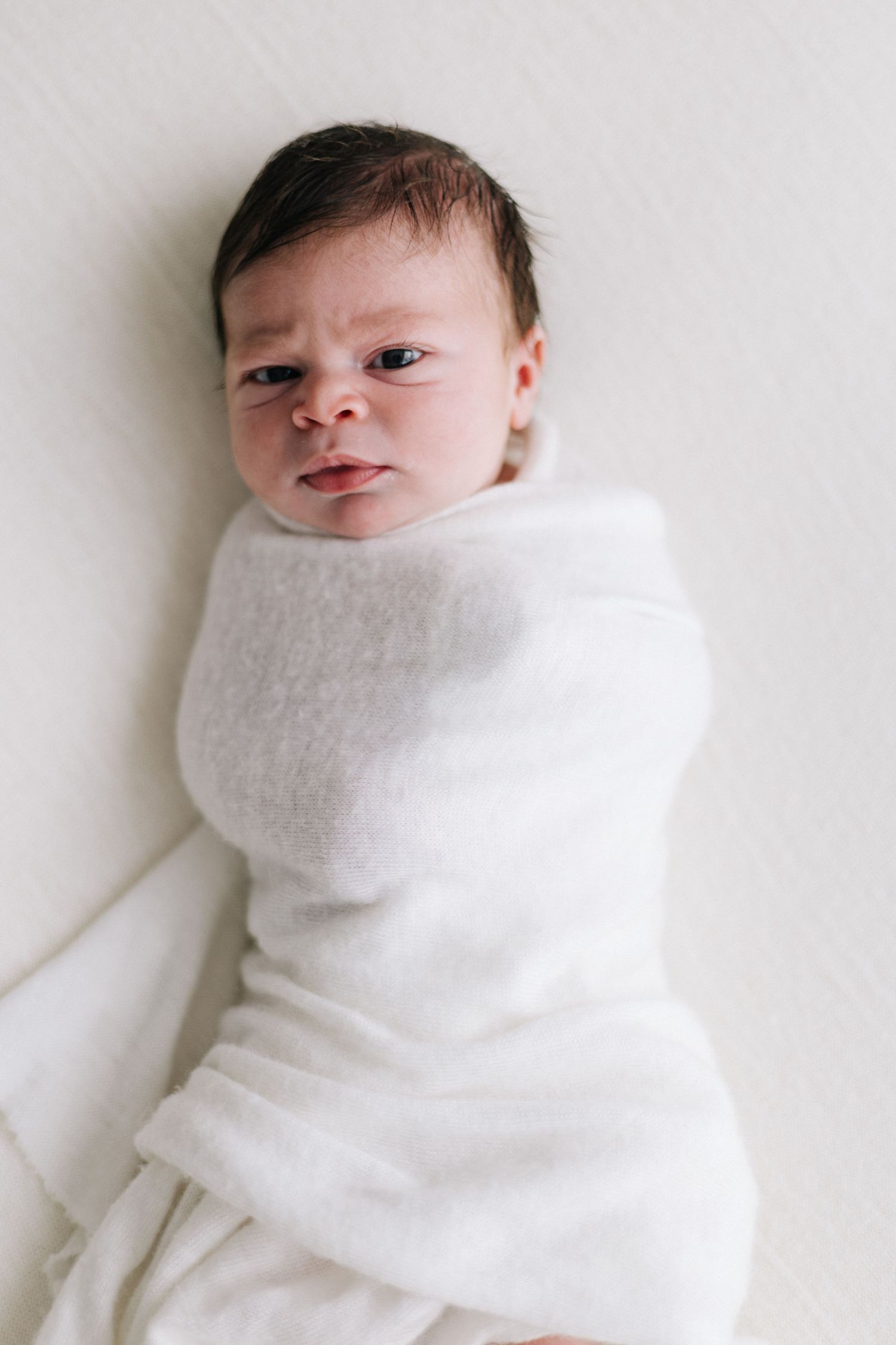 Awake newborn baby swaddled in white wrap lying on white background.