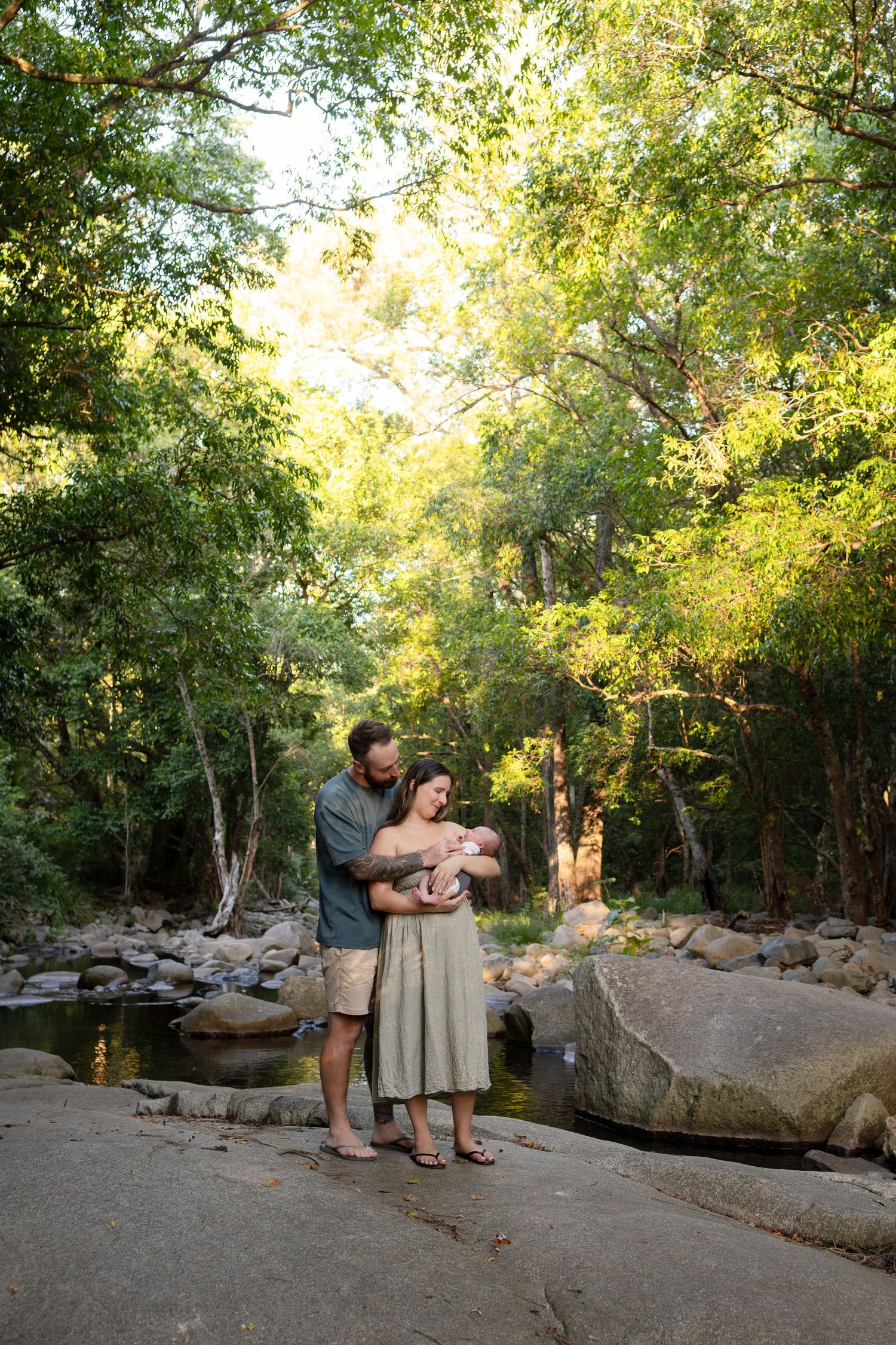A couple stands on rocky terrain by a creek in a lush forest, tenderly cradling their newborn baby.