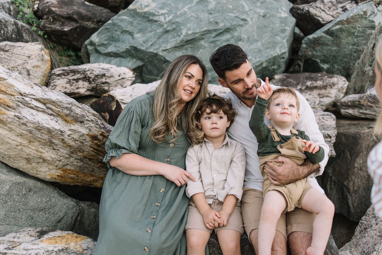 Family of four smiling together on rocky terrain, parents with two young children in natural outdoor setting.