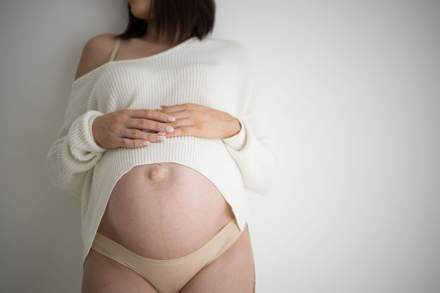 Pregnant woman in white sweater and beige underwear cradling her bare baby bump against a white wall.