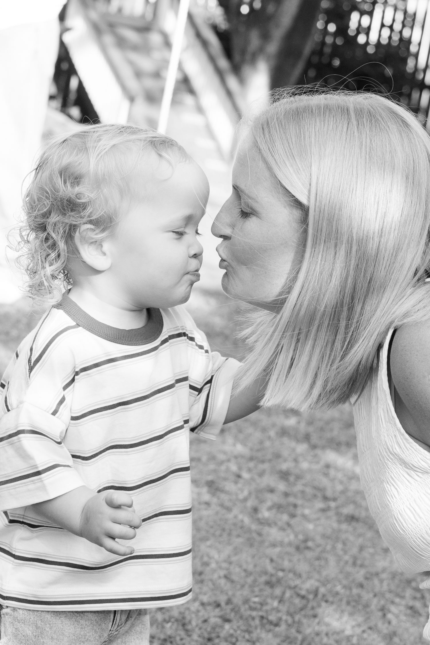 Black and white photo of a young mother leaning in to kiss her curly-haired toddler outdoors.