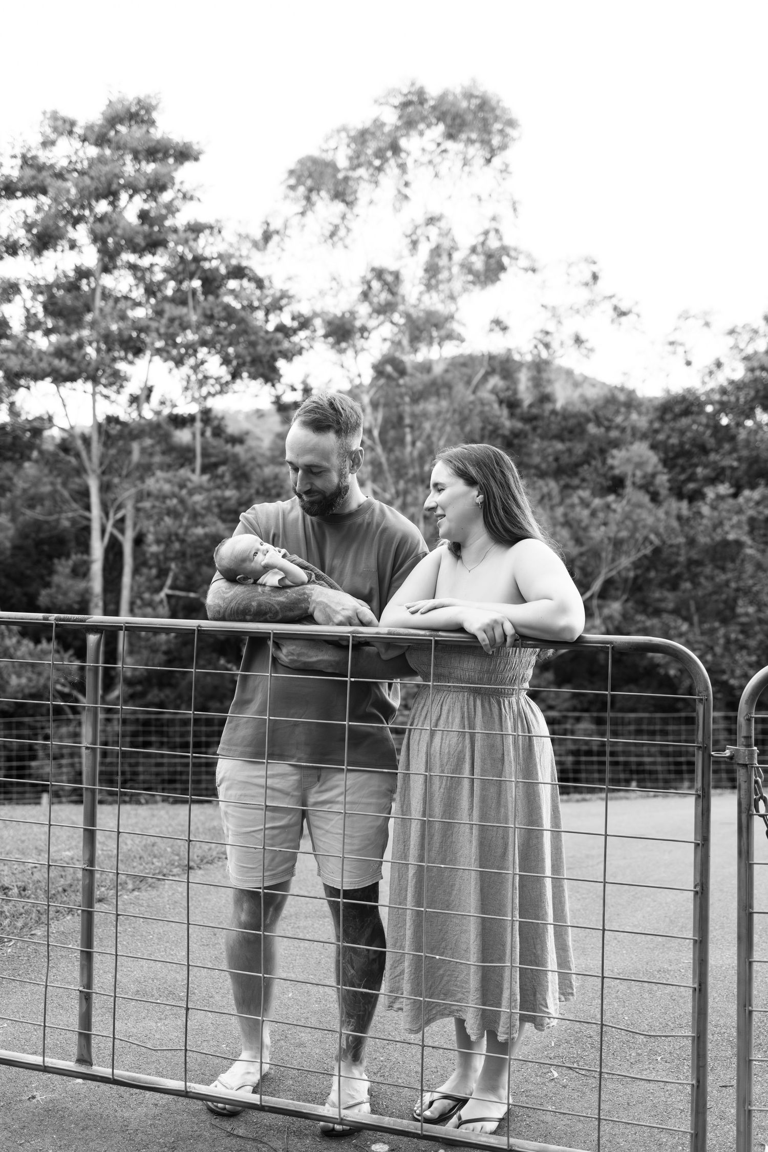 A couple stands at a farm gate in black and white, the father holding their newborn baby outdoors.