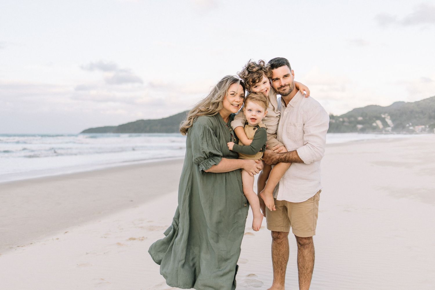 Happy family of four smiling together on a sandy beach at sunset, parents holding young children.