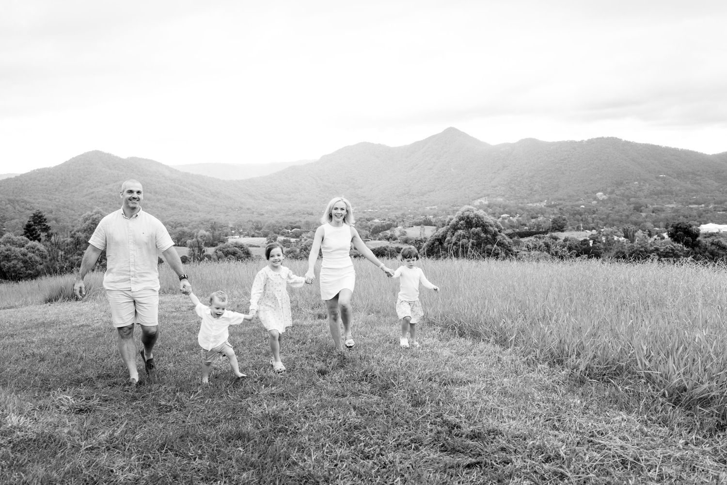 Black and white photo of a family of four holding hands walking through a green field with mountains in the background.