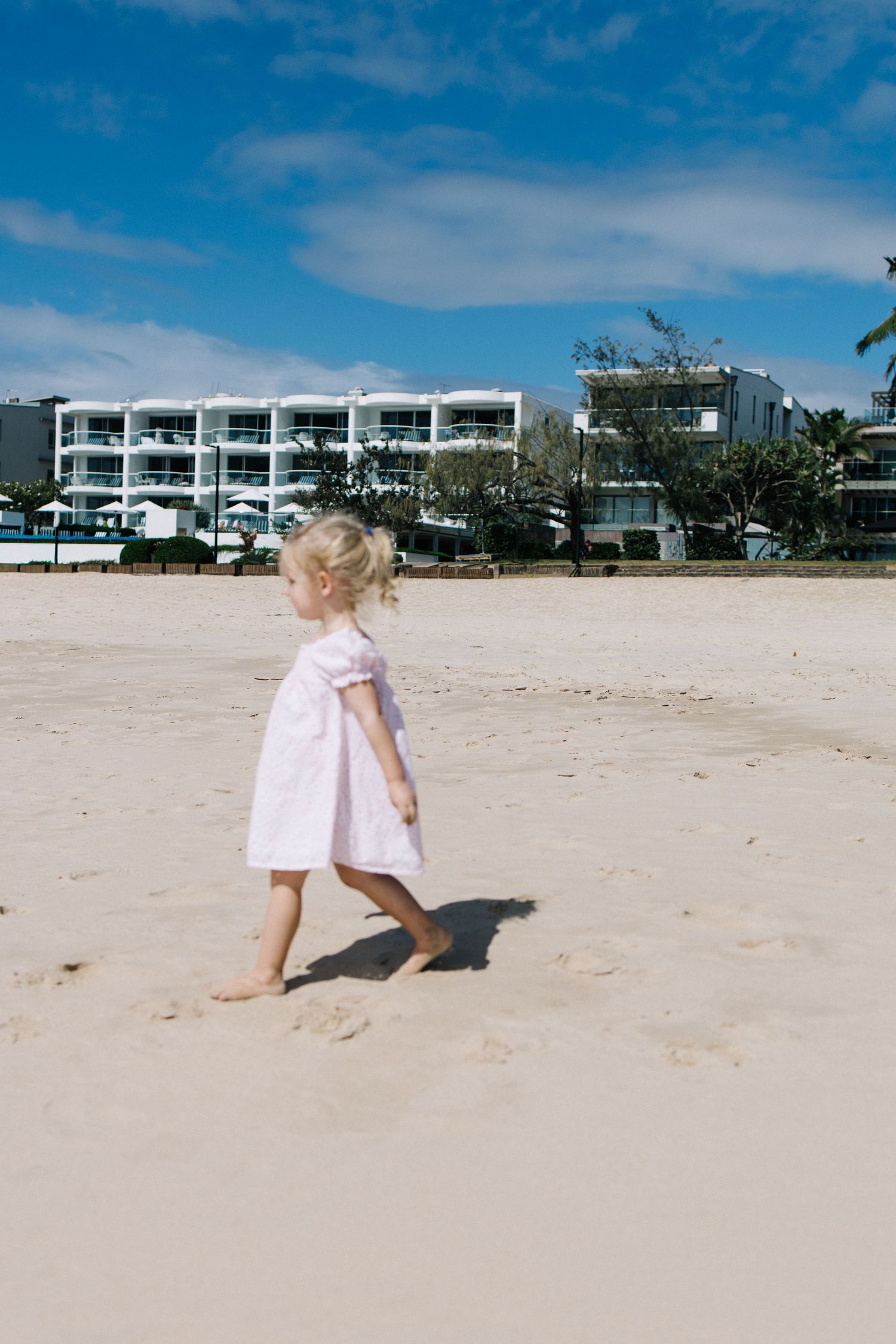 A toddler girl in a pink dress walks barefoot on a sandy beach with a resort building and blue sky behind her.
