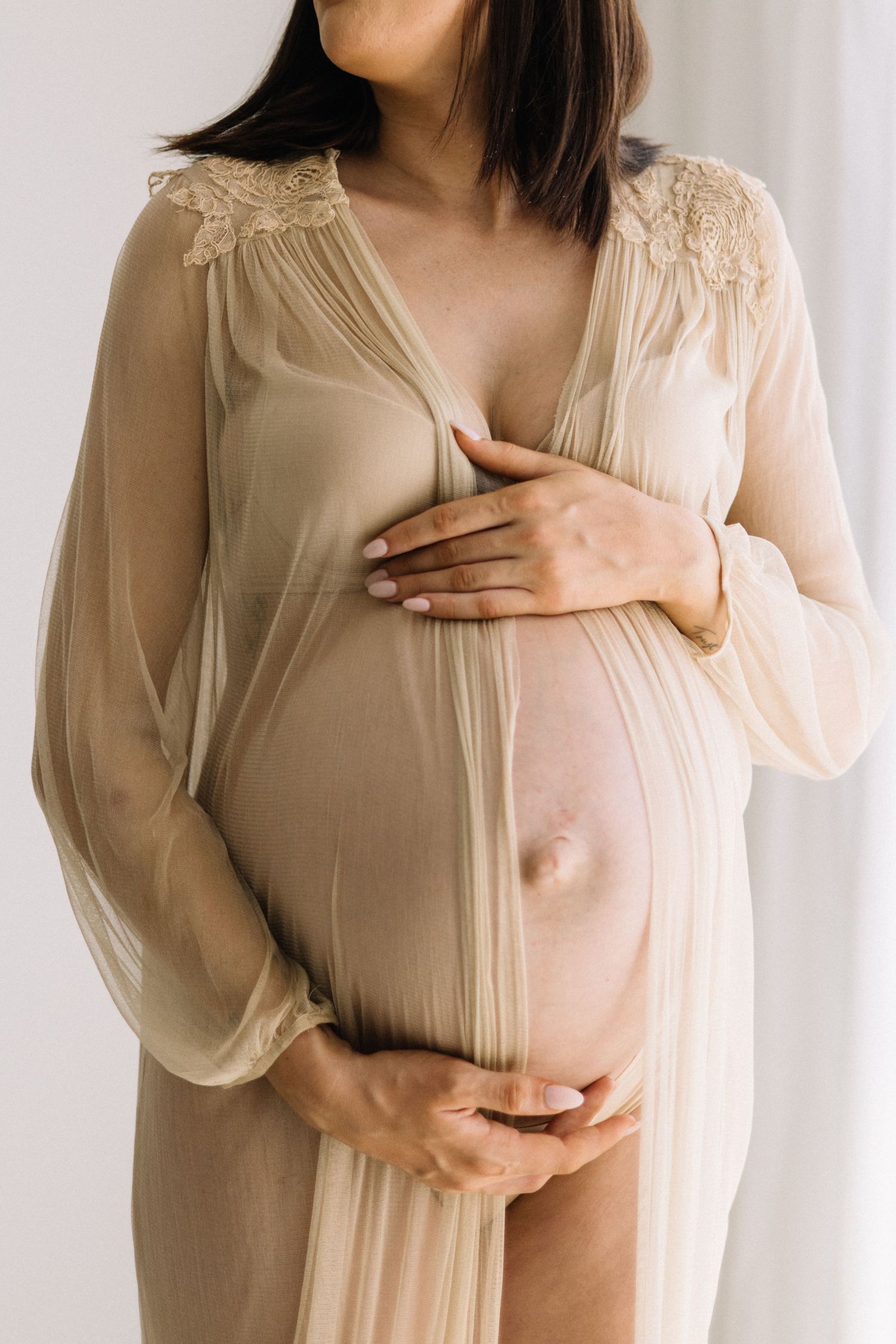 Pregnant woman in sheer beige lace robe cradling her bare baby bump with both hands.
