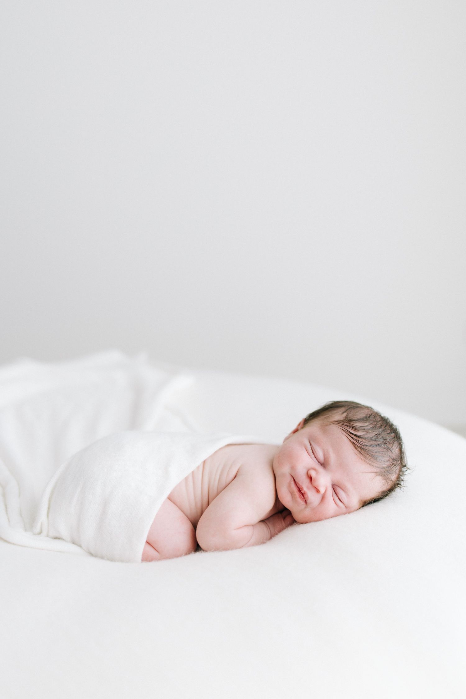 Sleeping newborn baby lying on white blanket, smiling softly with dark hair, serene nursery setting.