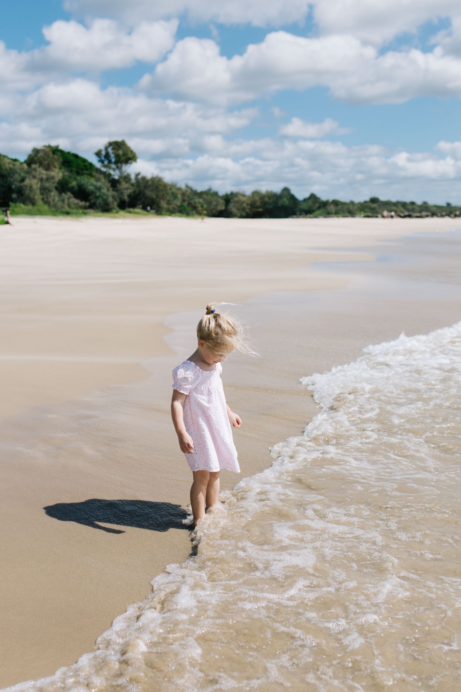 Young girl in pink dress walking along sandy beach shoreline with gentle waves, blue sky and green trees in background.