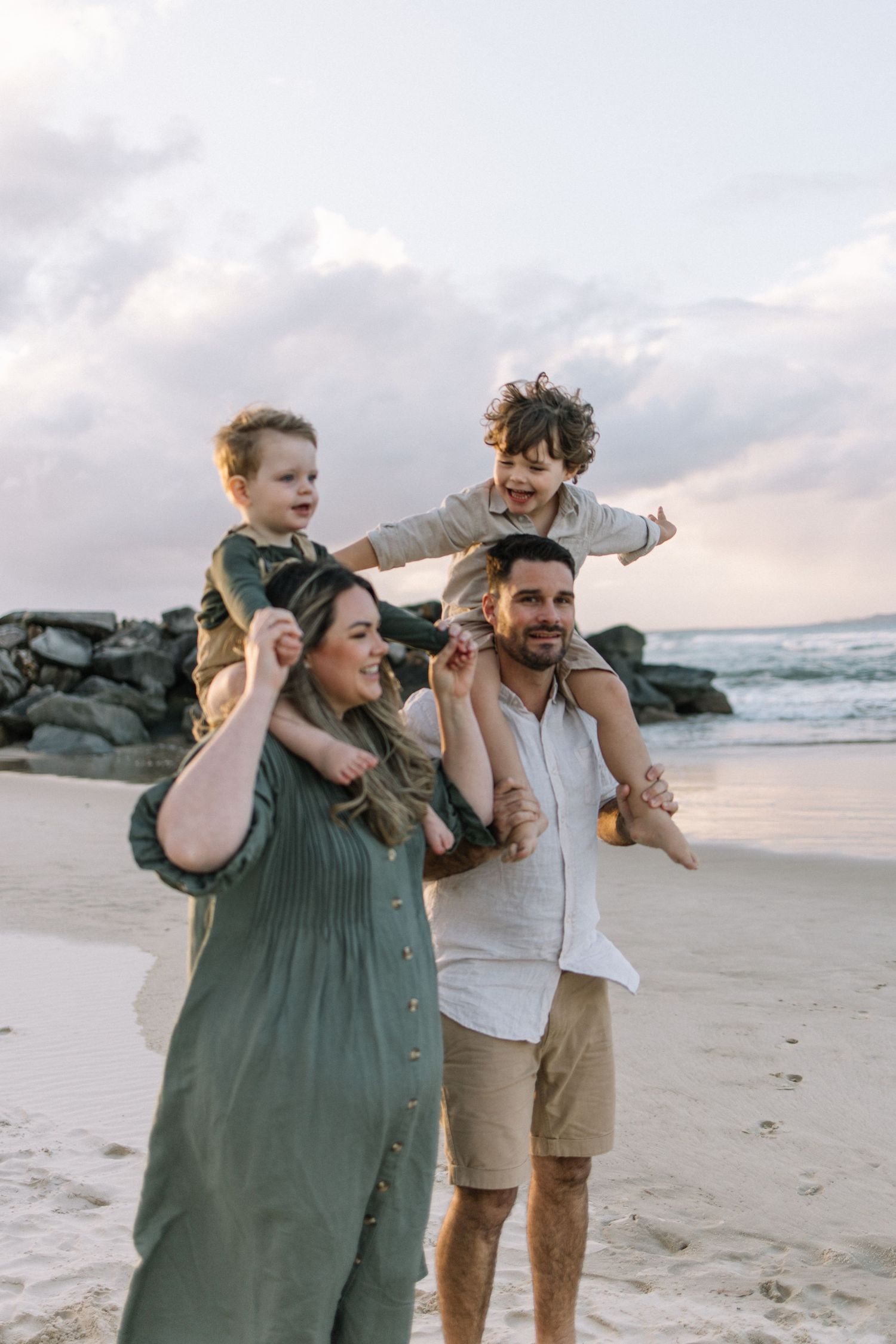 Family of four enjoying a beach day, with two children riding on parents shoulders near rocky shoreline at sunset.