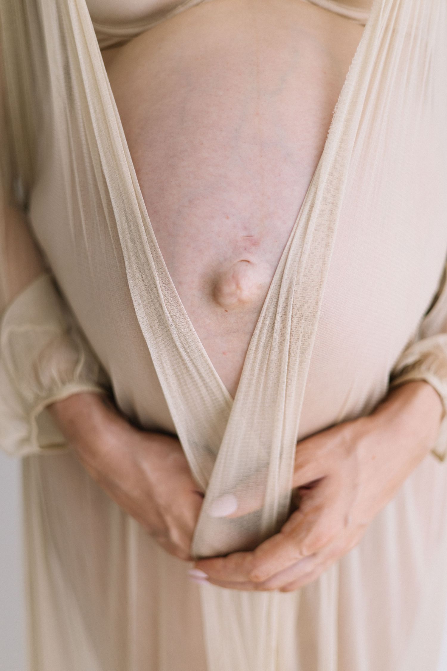 Close-up of pregnant belly with protruding navel held by hands in beige sheer fabric dress.
