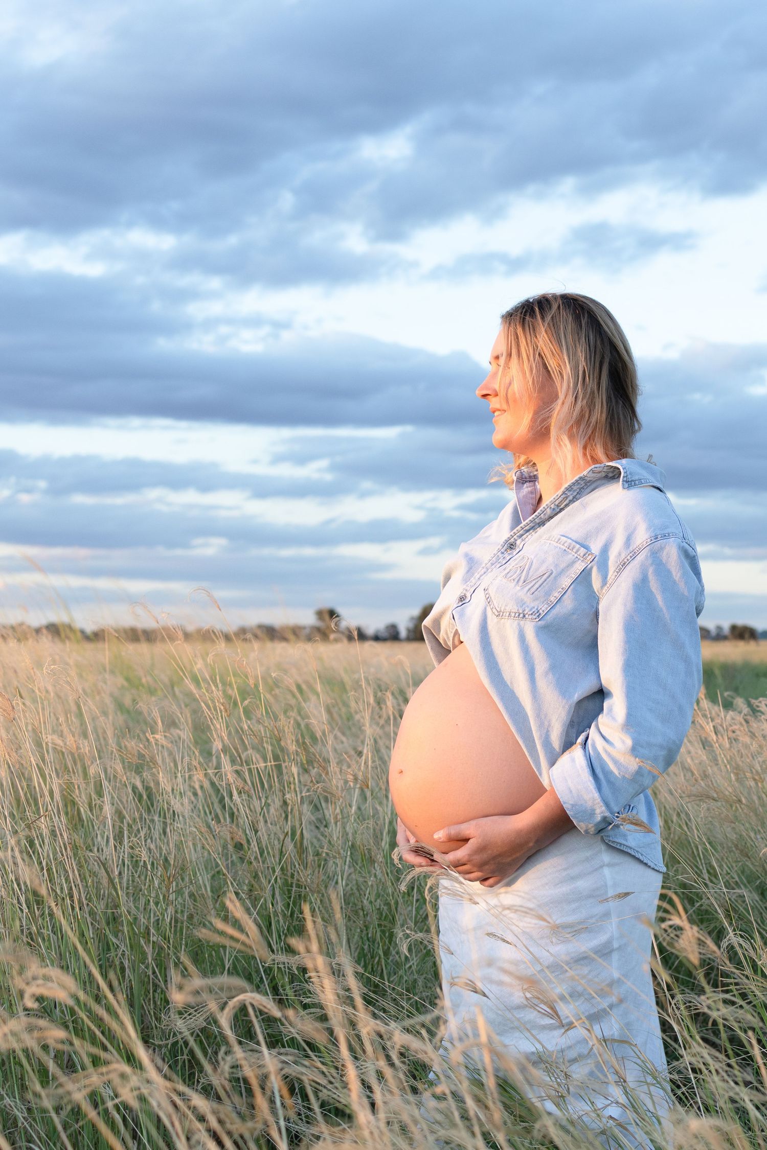 Smiling pregnant woman holding belly in tall grass field at golden sunset.