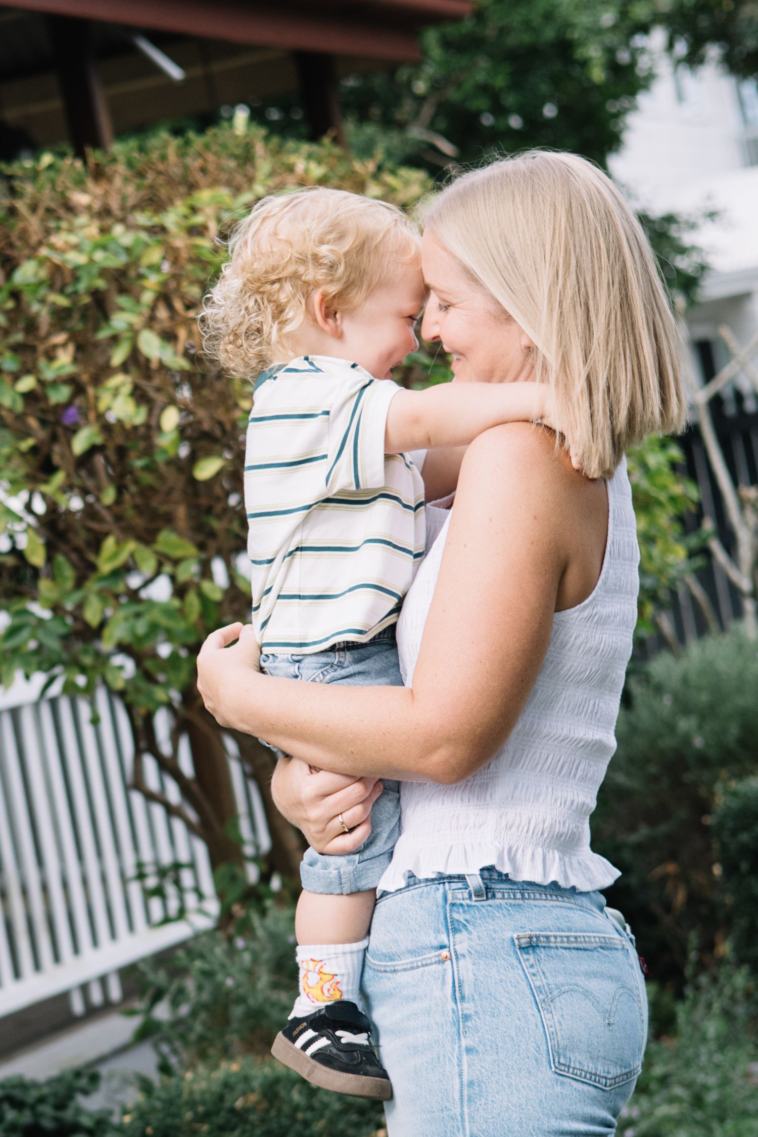A blonde woman in a white tank top and jeans holds a curly-haired toddler, touching noses affectionately outdoors.