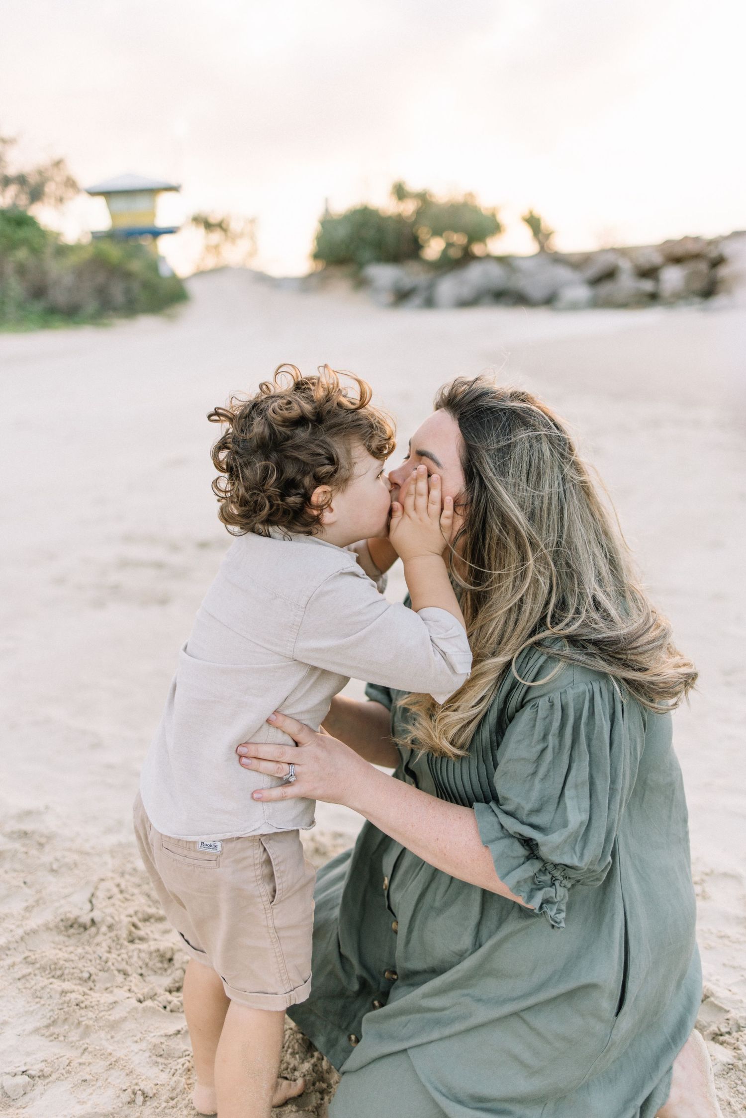 A mother crouches on the beach as her curly-haired toddler gives her a sweet kiss.