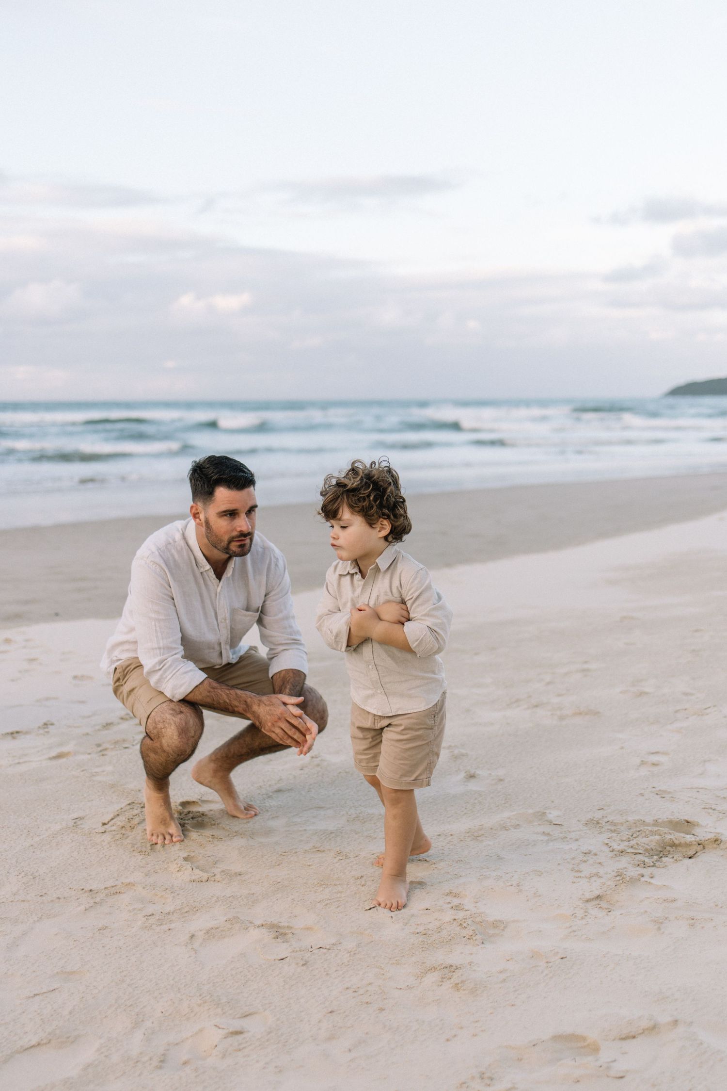 Father crouching beside young son on sandy beach at dusk, ocean waves in background.