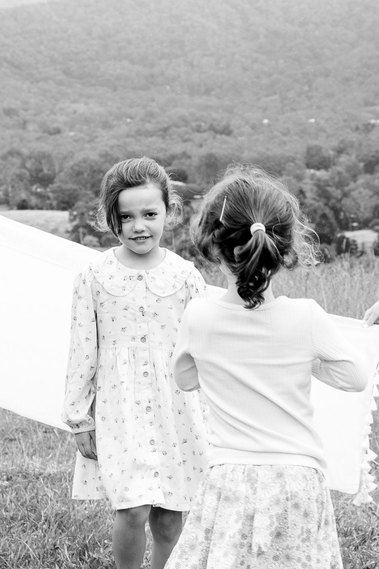 Two young girls in dresses hang laundry outdoors with green mountains in the background, black and white photo.