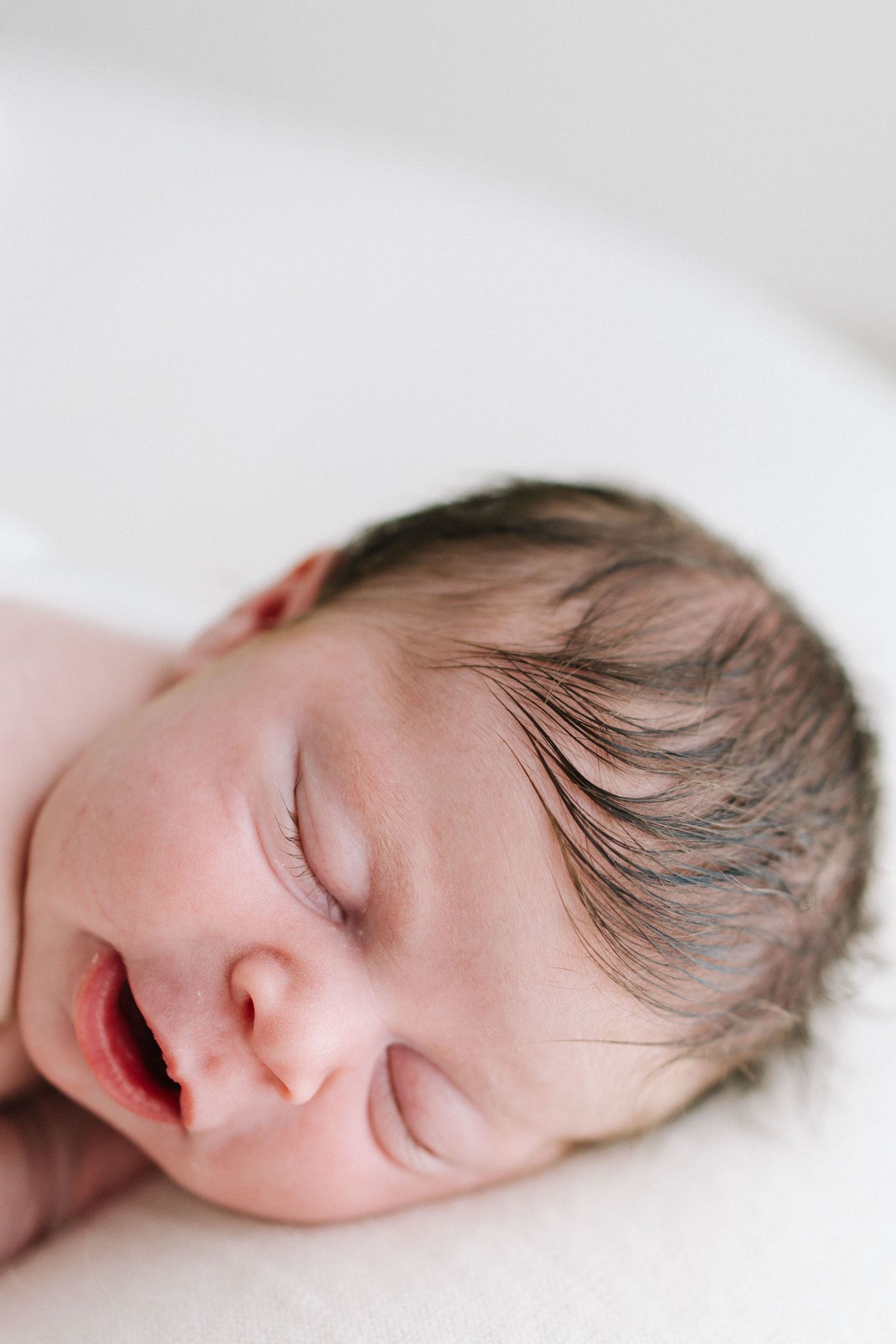 Peaceful sleeping newborn baby with dark hair, eyes closed, on a white surface.