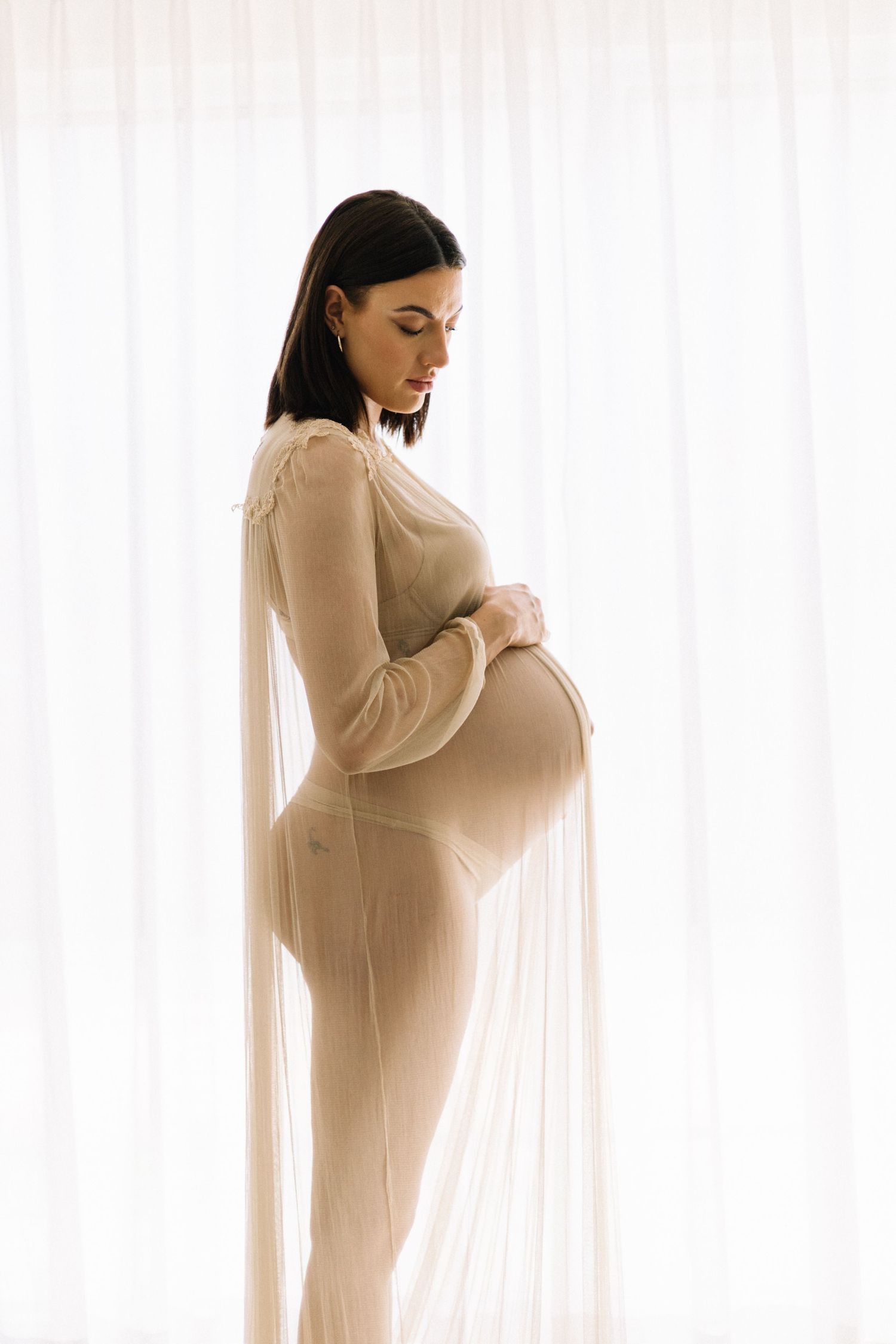 Pregnant woman in sheer beige draped fabric standing by white curtains cradling her belly.