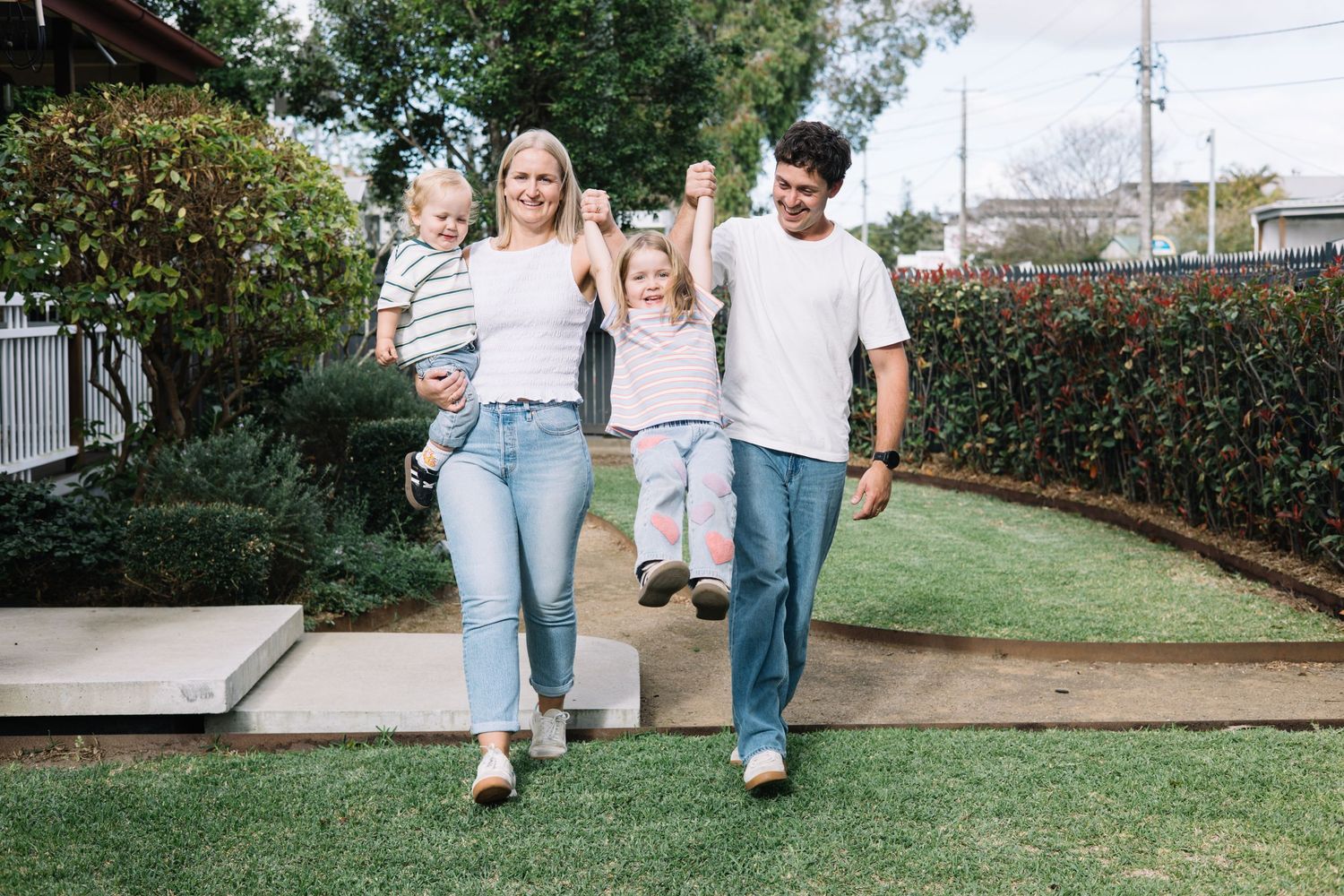 Happy family of four walking outdoors on green lawn, parents swinging children, wearing casual white and blue outfits.