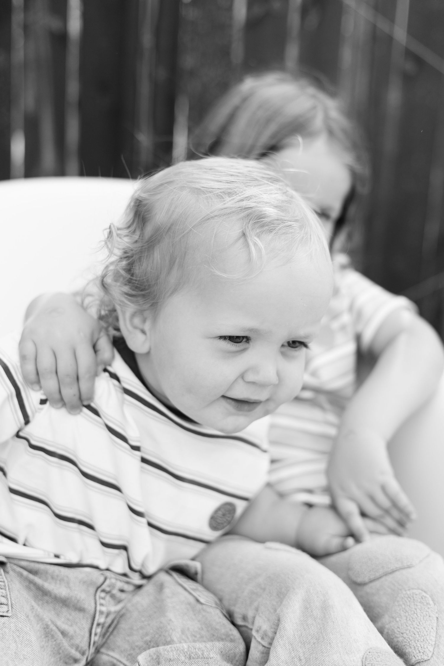 Black and white photo of a smiling toddler with curly hair sitting with an older child outdoors.