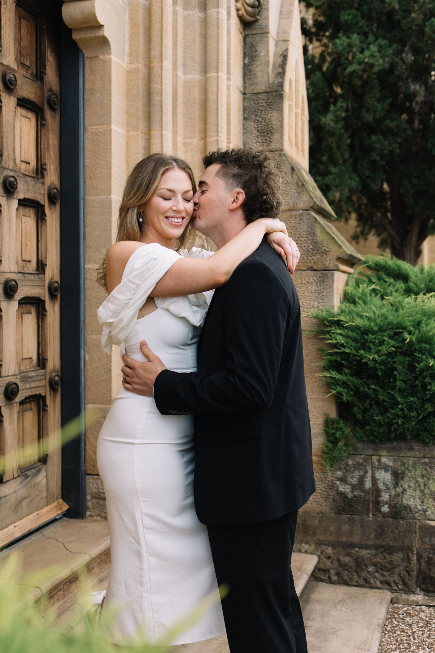 A couple embraces and shares a kiss outside a stone church, the woman in a white dress smiling joyfully.