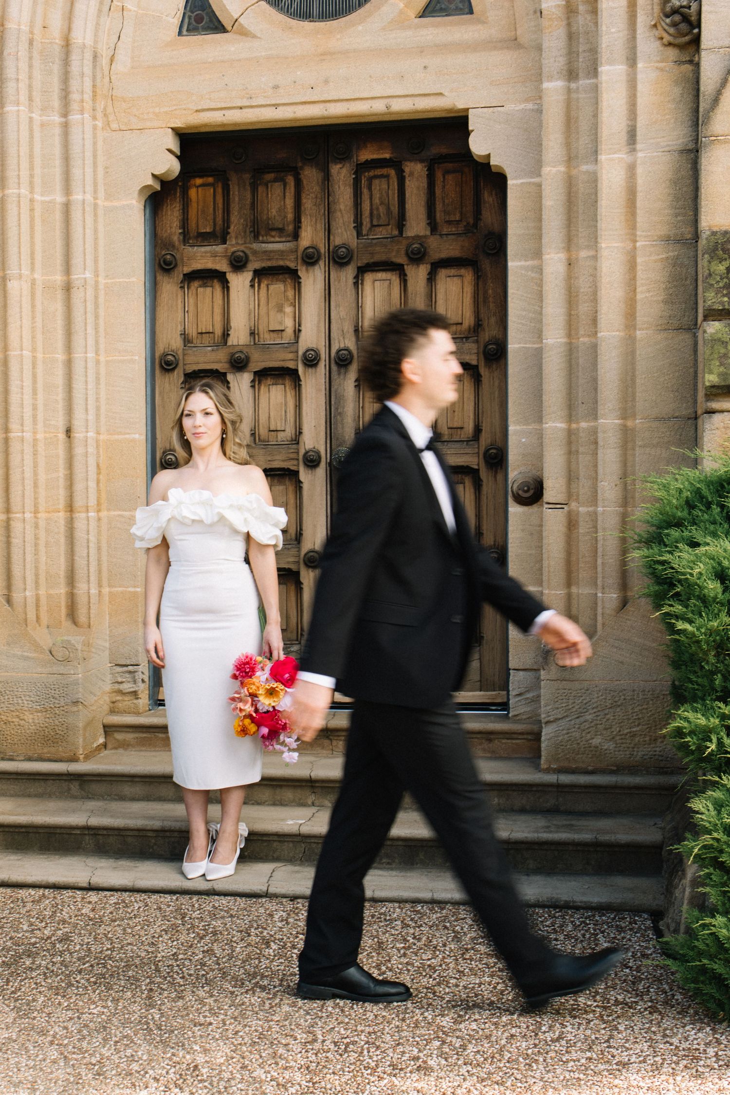Bride in white off-shoulder midi dress holds colorful bouquet by ornate wooden doors as groom in tuxedo walks past.